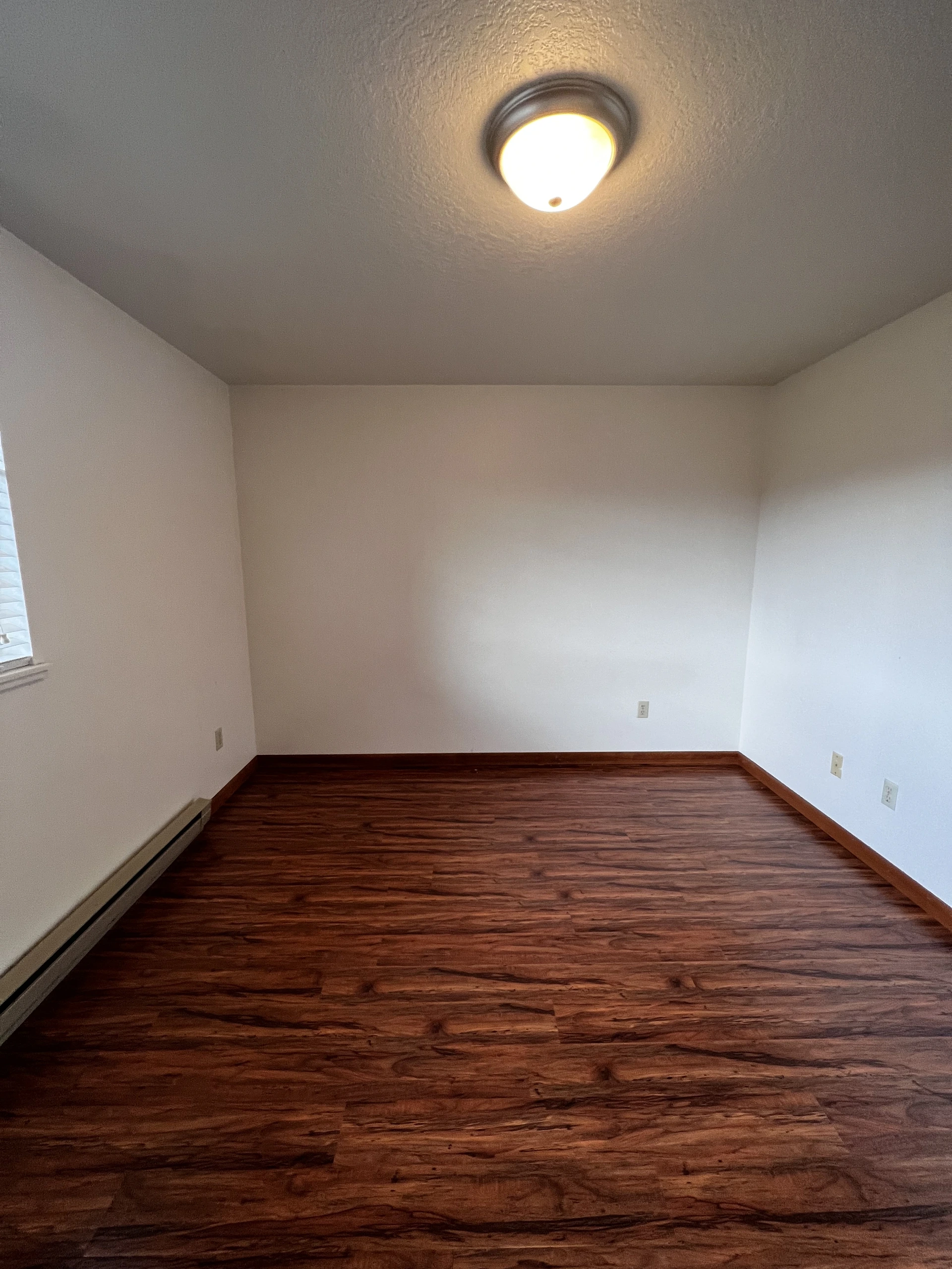 Empty room with dark wood flooring, light walls, a ceiling light, a window, and several wall outlets.