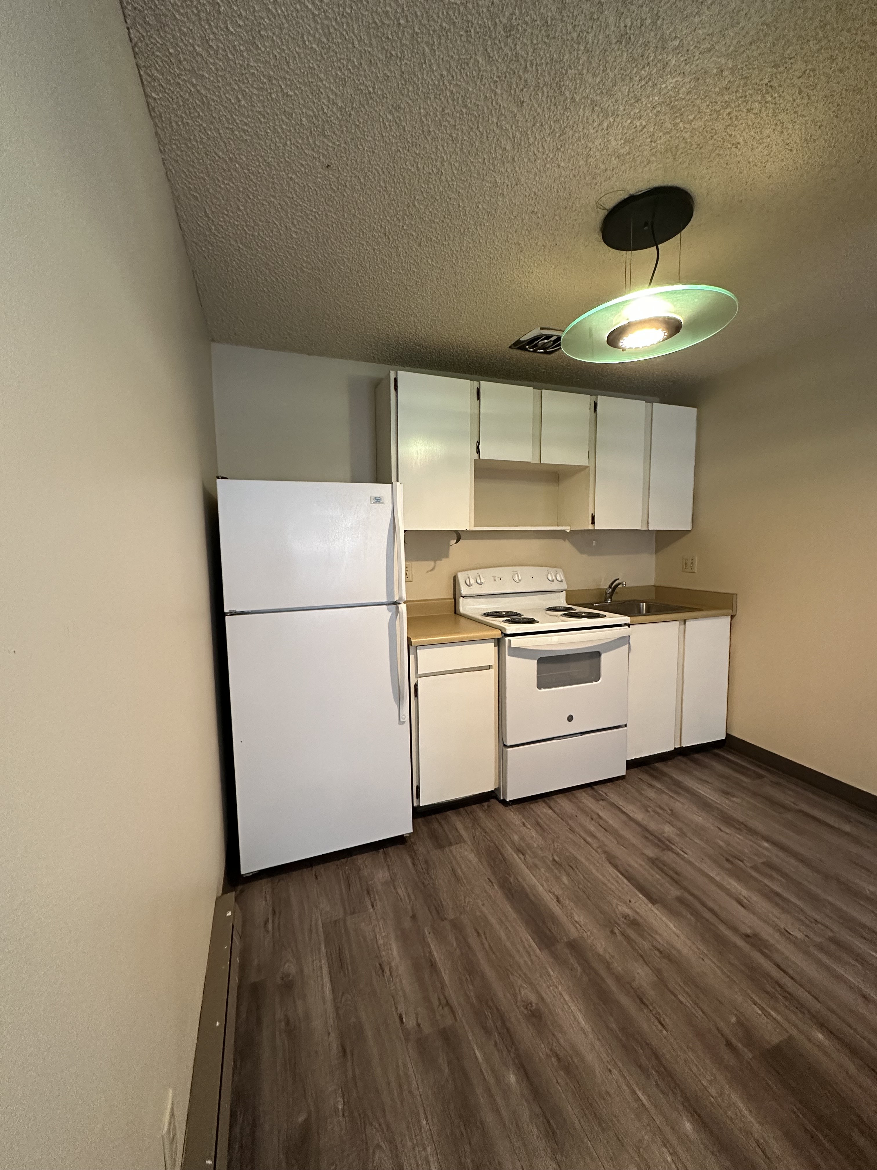 A small kitchen with a refrigerator, electric stove, sink, white cabinets, and a hanging ceiling light.