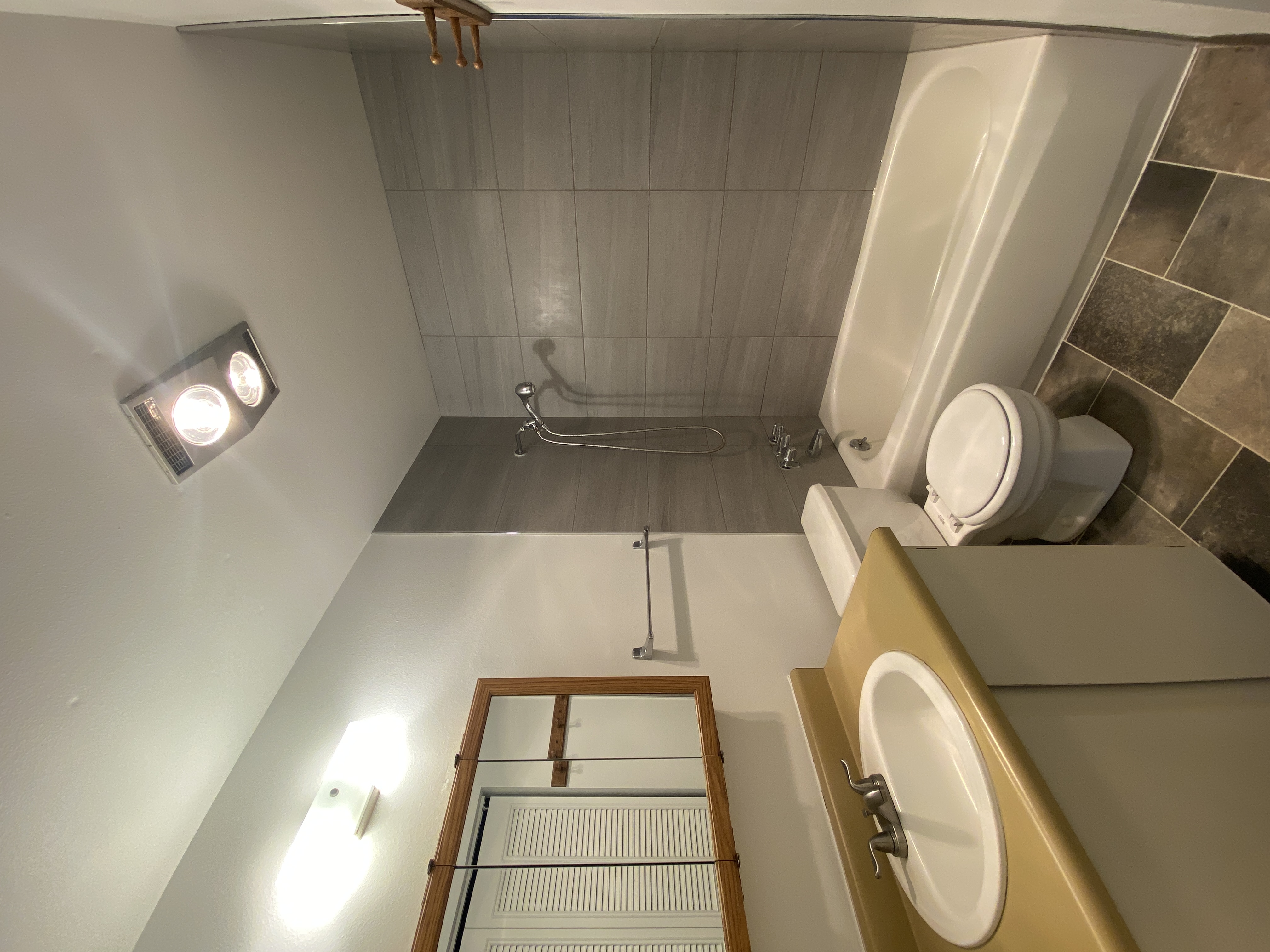 A bathroom with a sink and wood-framed mirror, a toilet, and a bathtub with a handheld shower against gray tile.