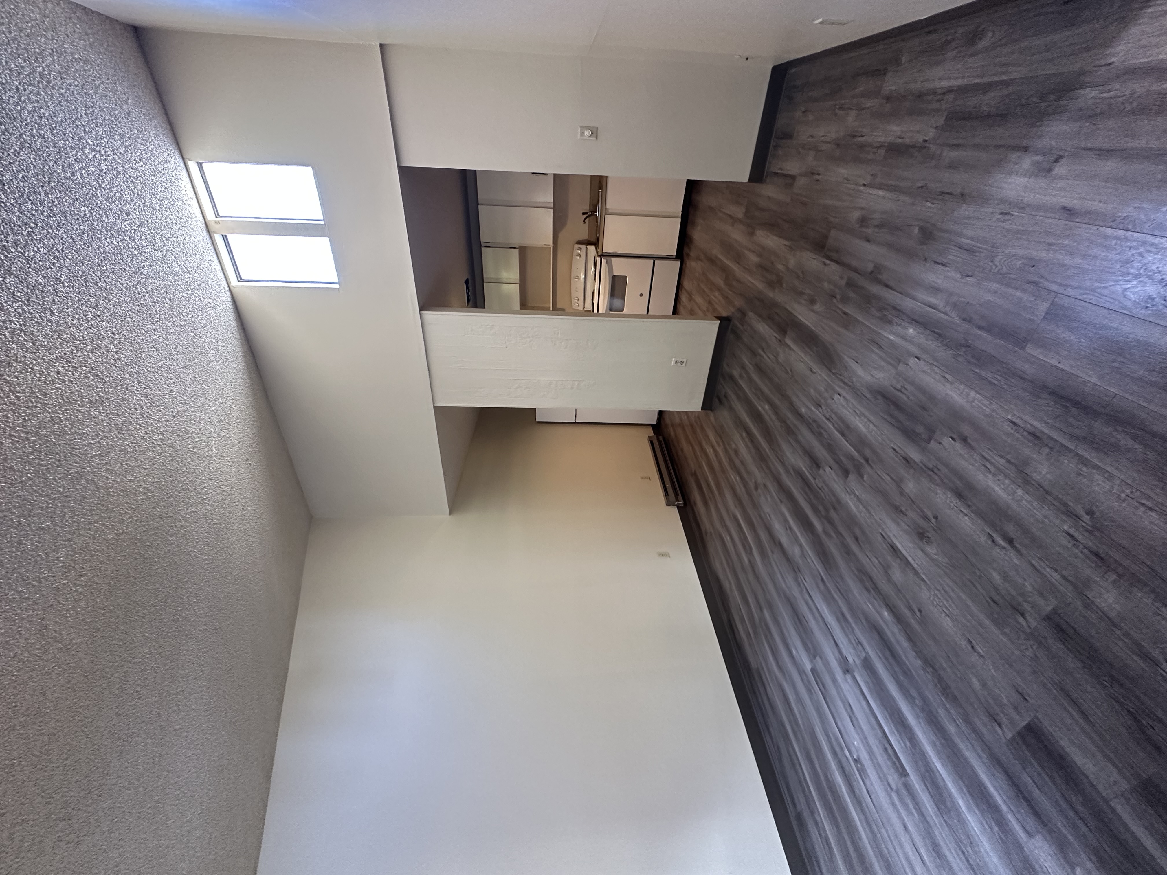 An empty room with dark wood-pattern flooring, a textured ceiling, and a kitchen visible through an opening.