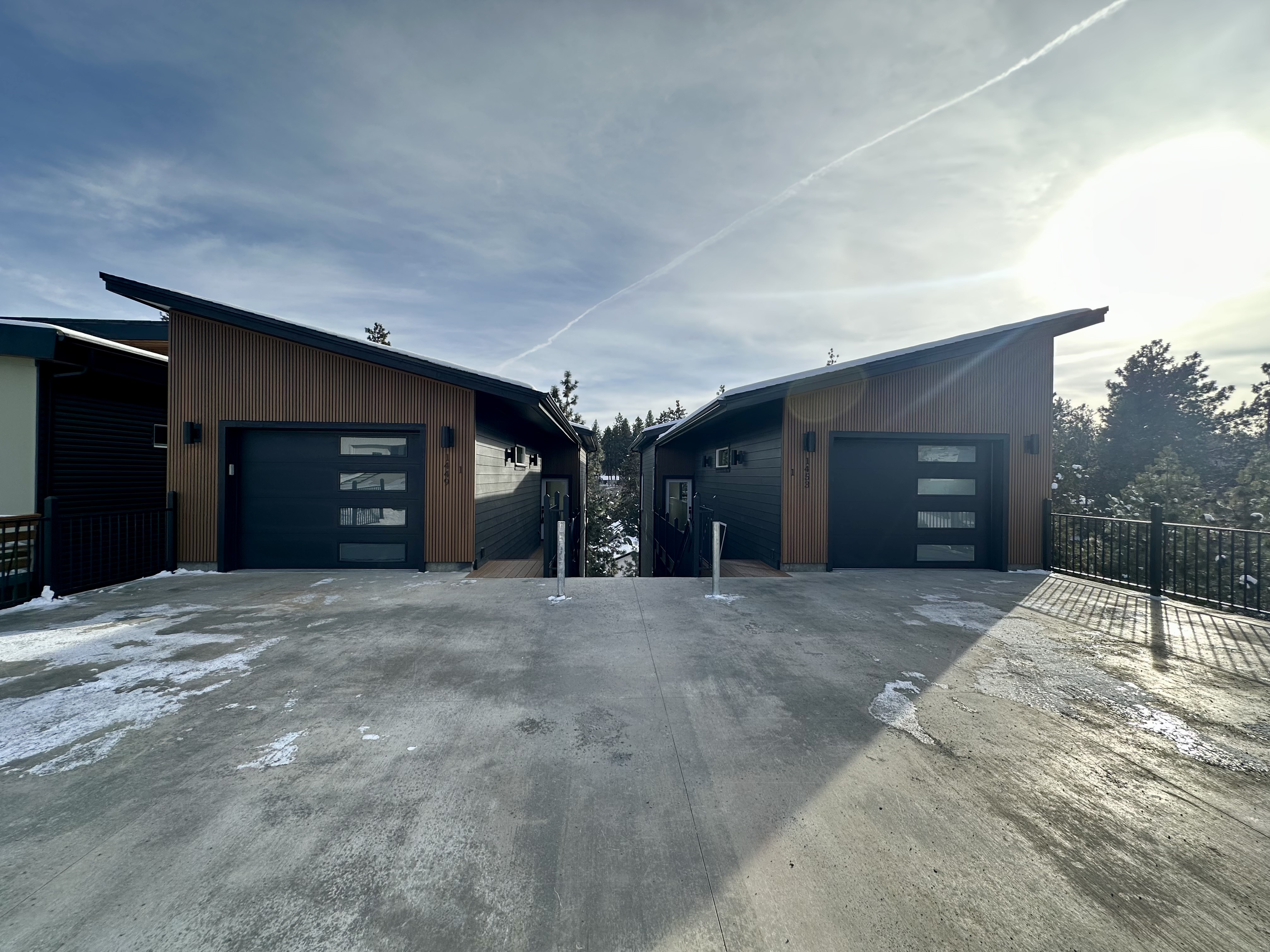 Modern dark-sided buildings with sloped roofs, doors, windows, railings, patches of snow, trees, and a partly cloudy sky.