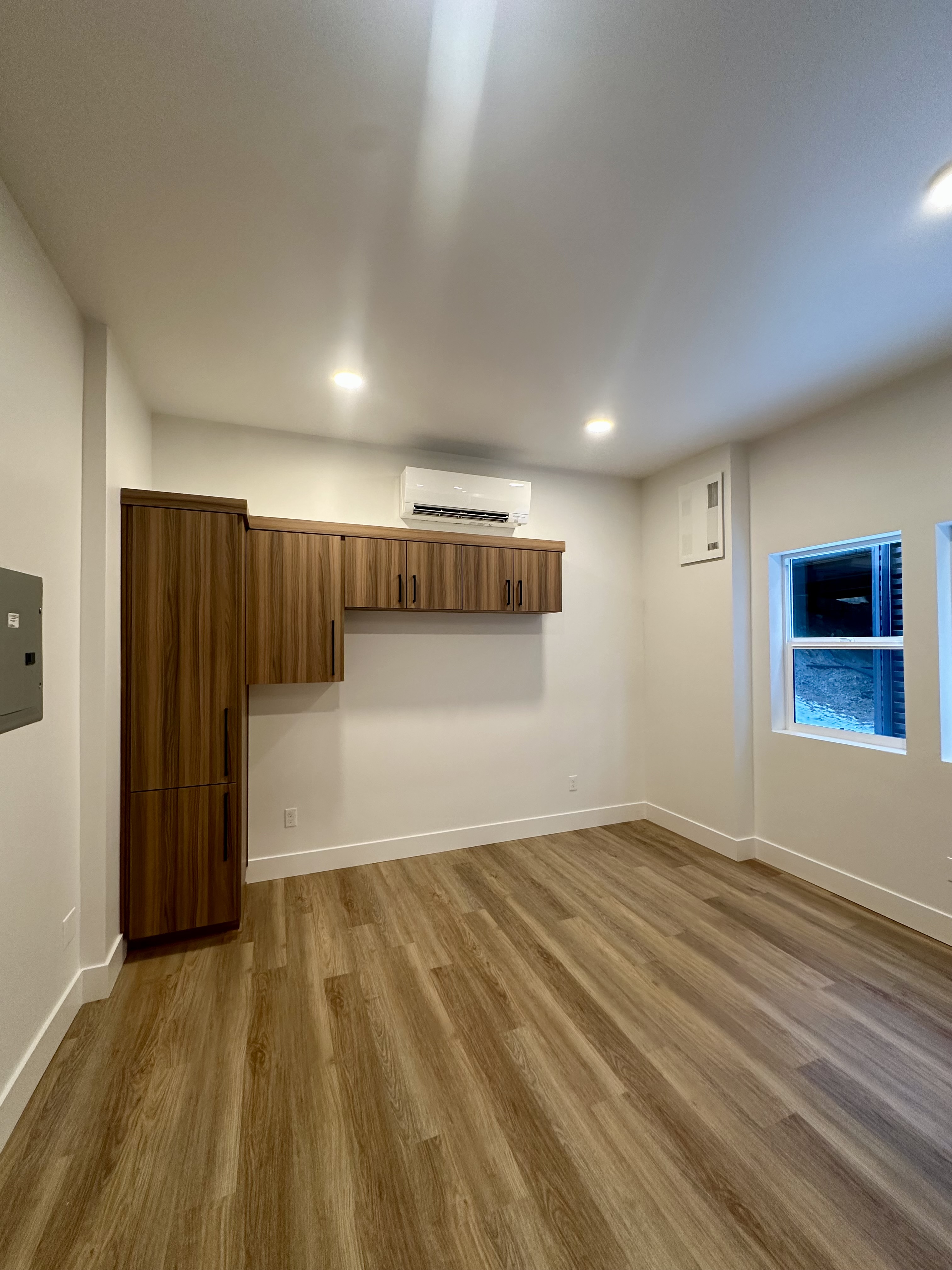 Empty room with wood cabinets, a wall-mounted air conditioner, recessed ceiling lights, wood-look flooring, and two windows.