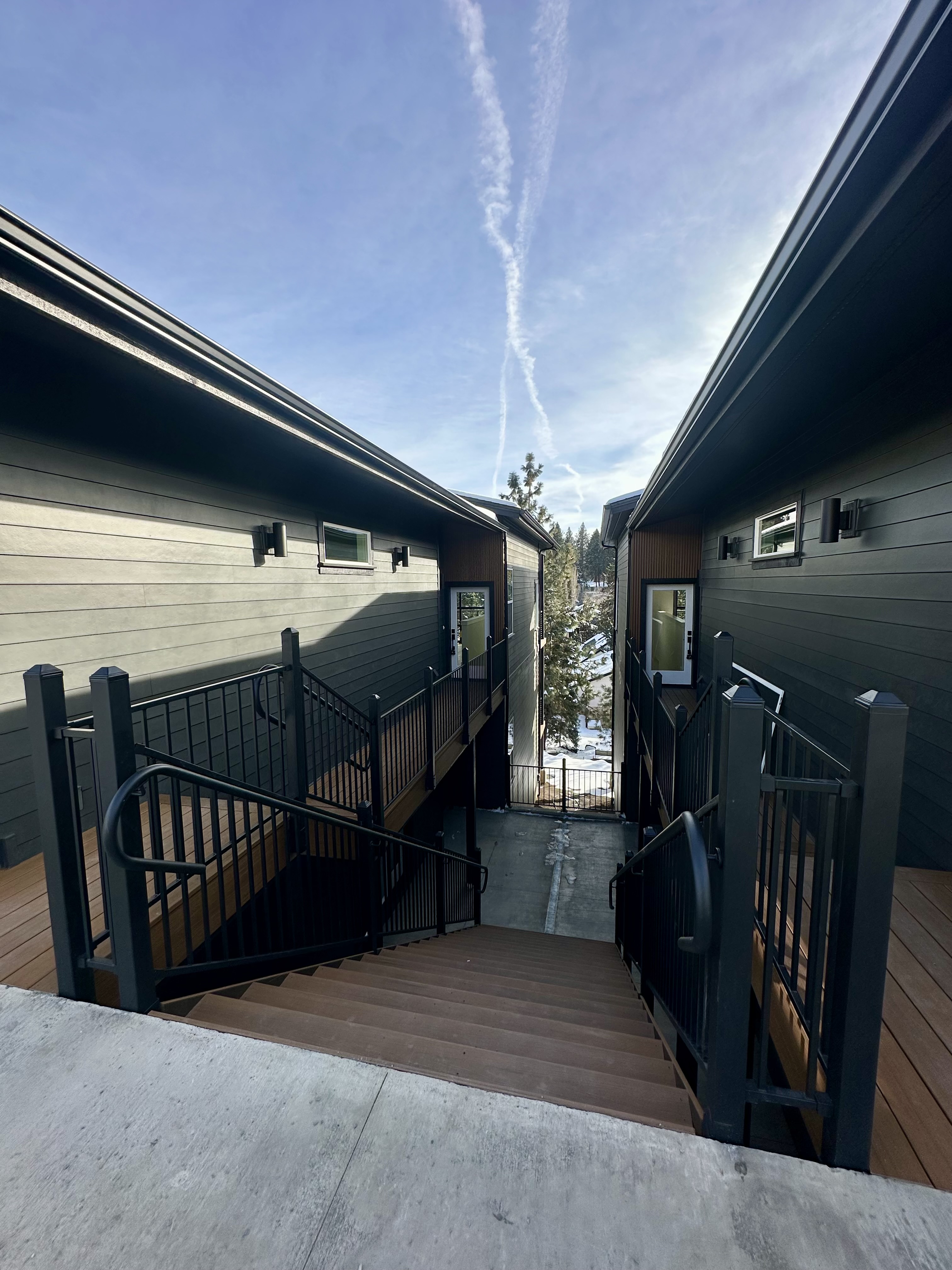 Exterior walkway with stairs and black railings between two buildings under a blue sky.