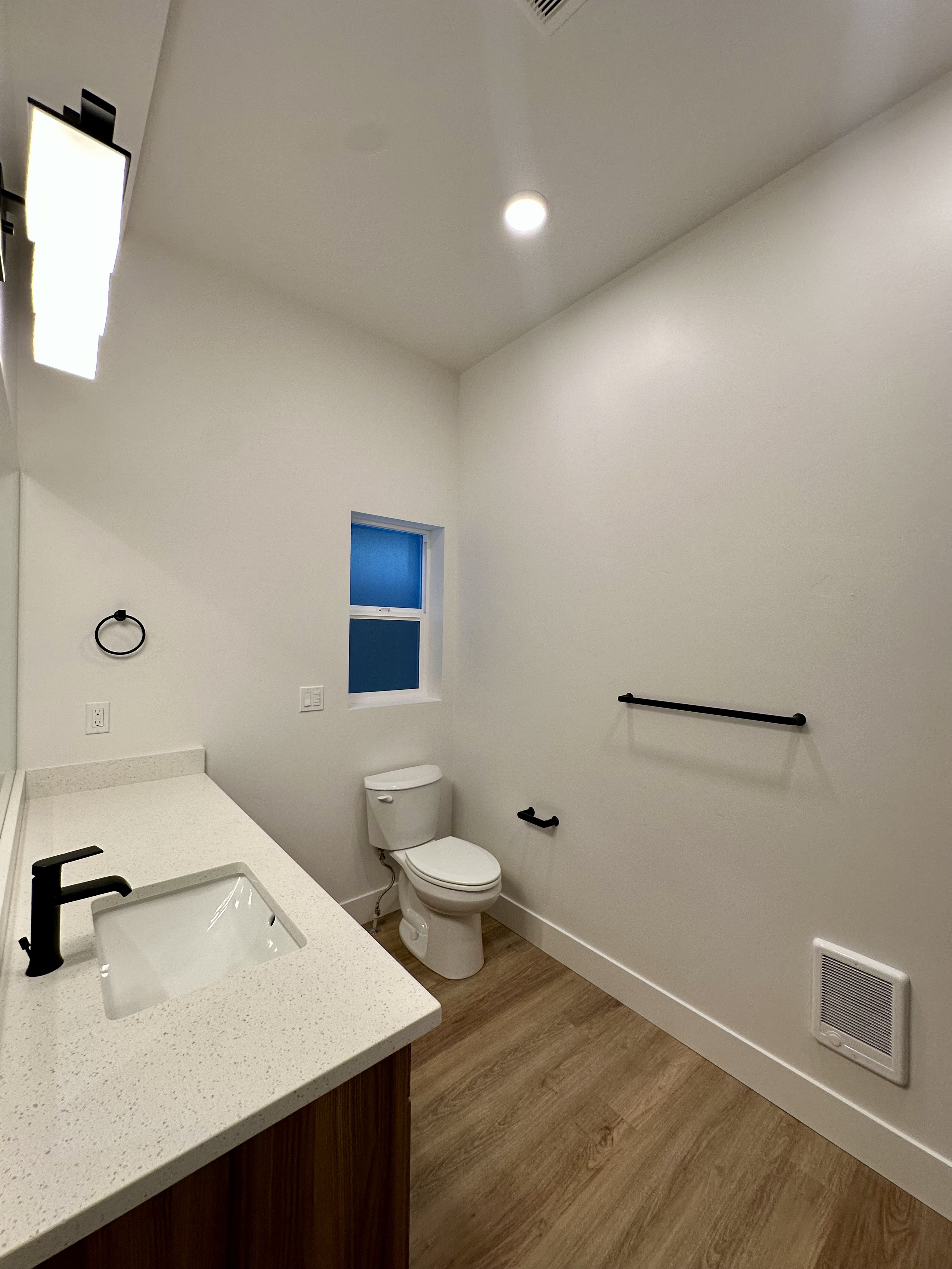 Bathroom with a sink vanity, toilet, narrow window, wall-mounted grab bars, and wood-look flooring.