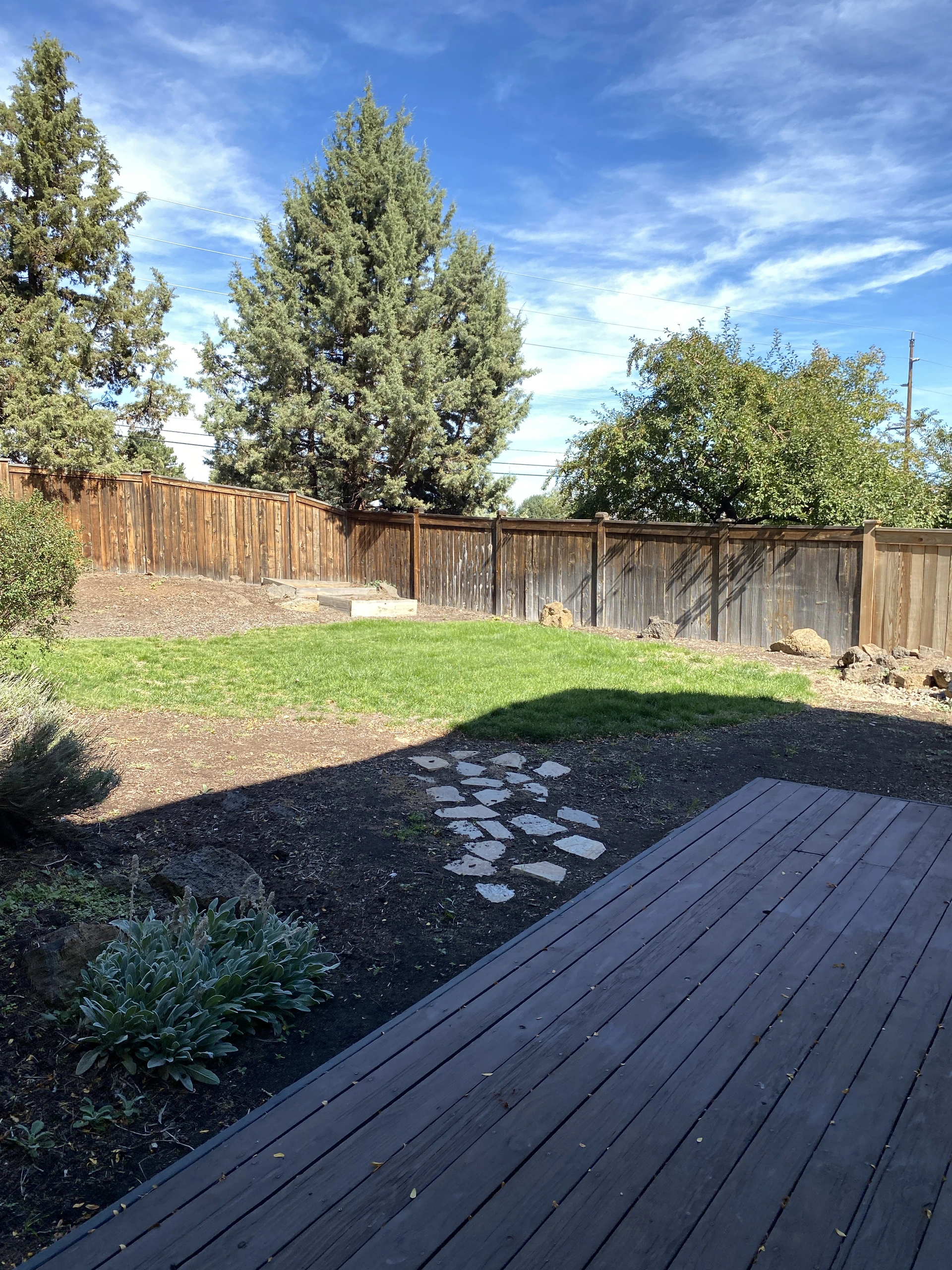 A wooden deck overlooks a fenced yard with grass, stepping stones, shrubs, and tall trees beneath a blue sky.