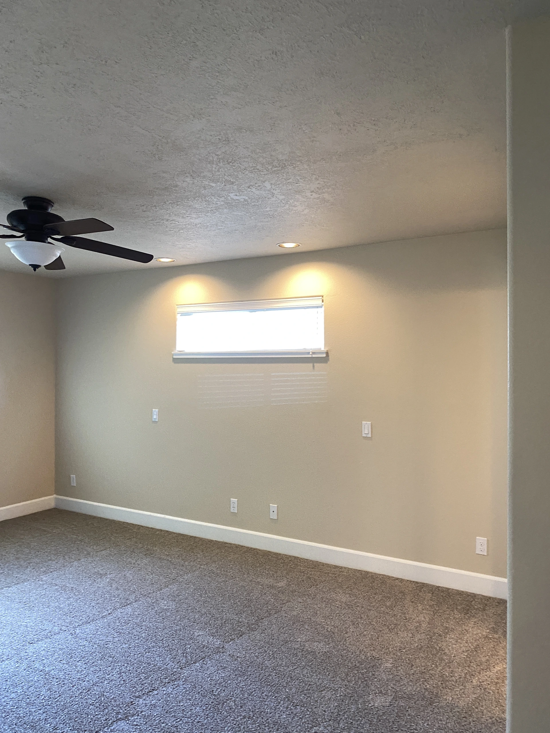 An empty room with gray carpet, beige walls, a high window, recessed ceiling lights, and a dark ceiling fan.