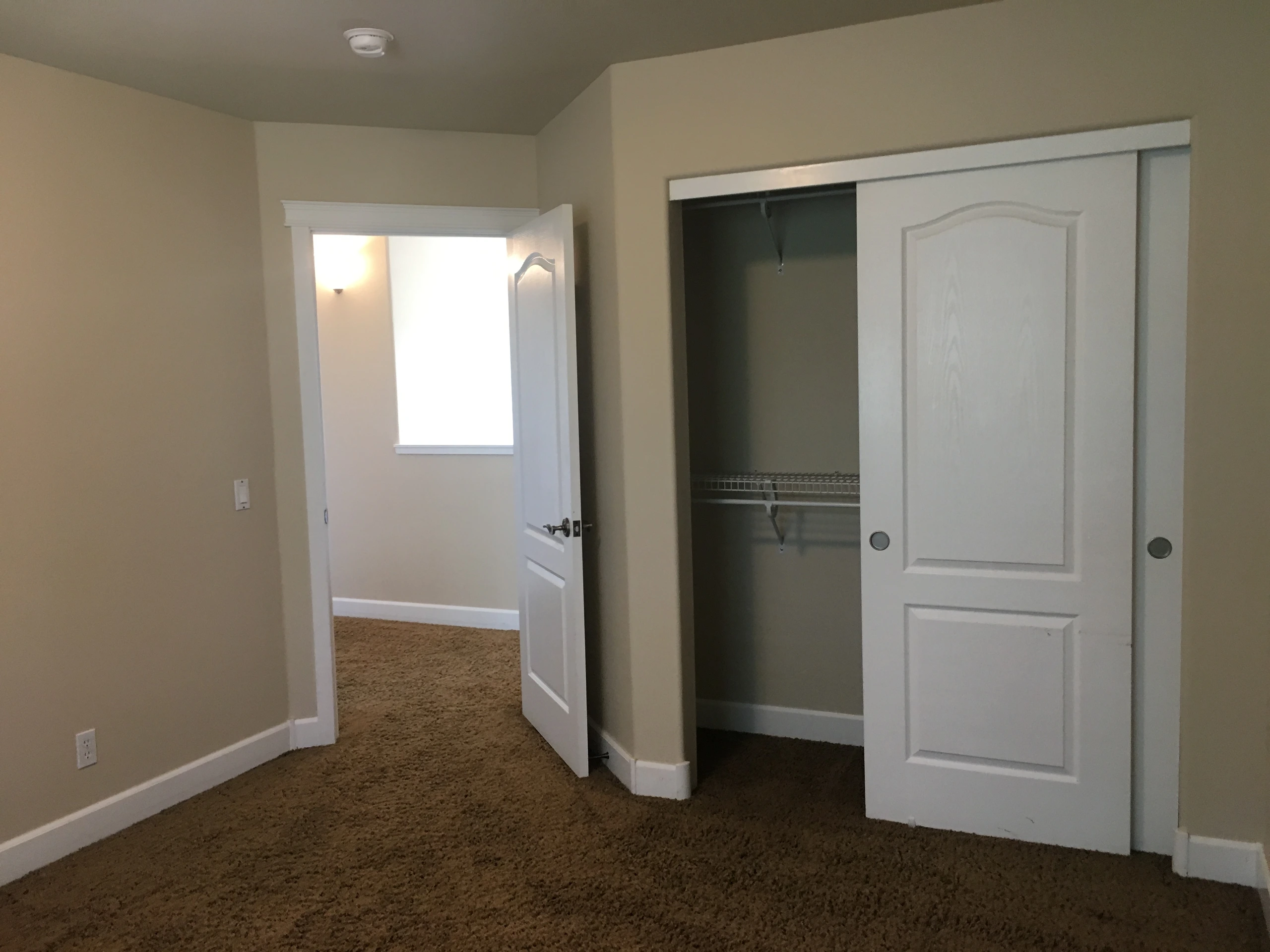 A carpeted room has beige walls, an open doorway, and a closet with sliding doors, one open to reveal a wire shelf.