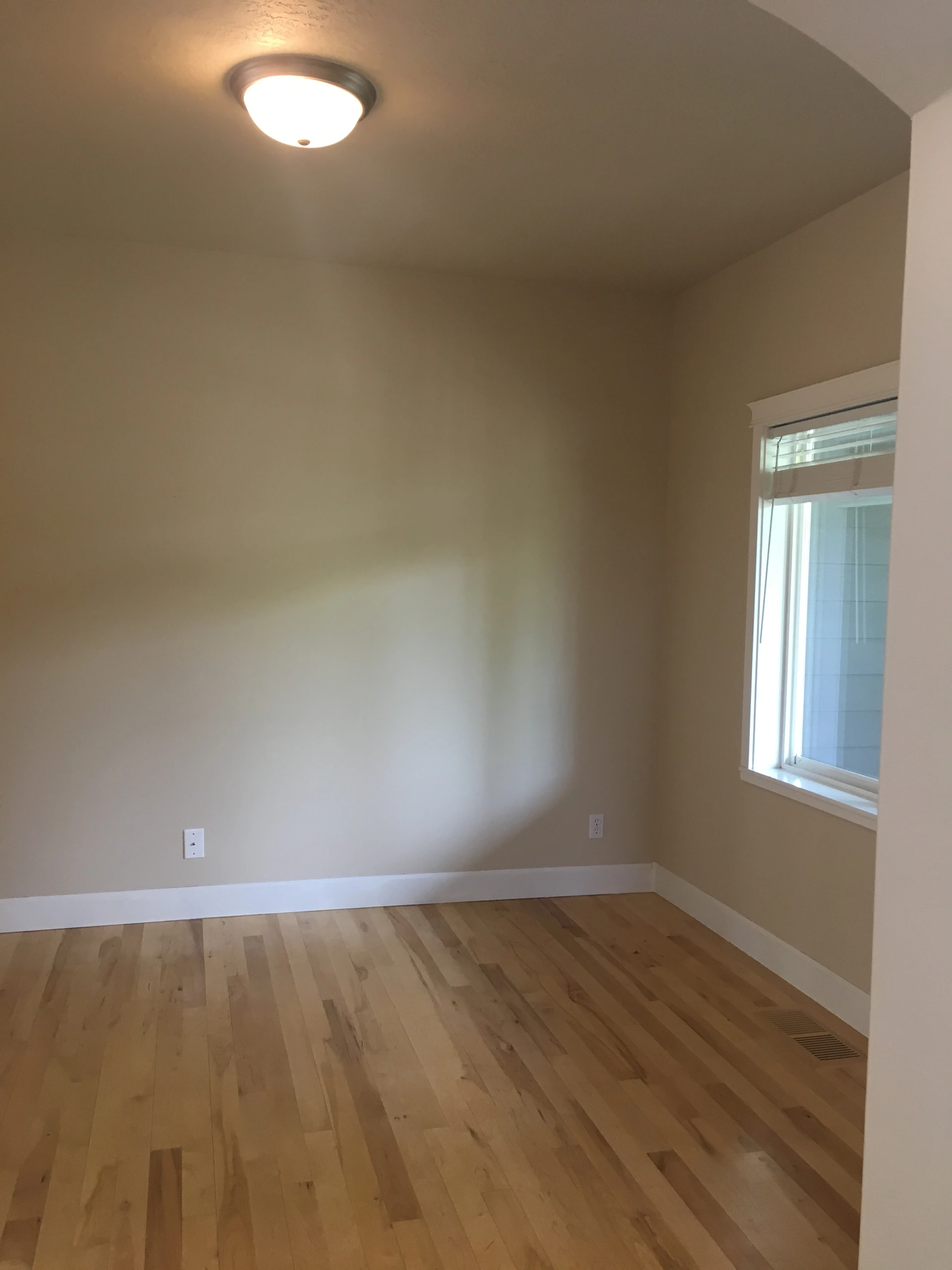 An empty room with beige walls, wood plank flooring, a ceiling light, and a window with blinds.