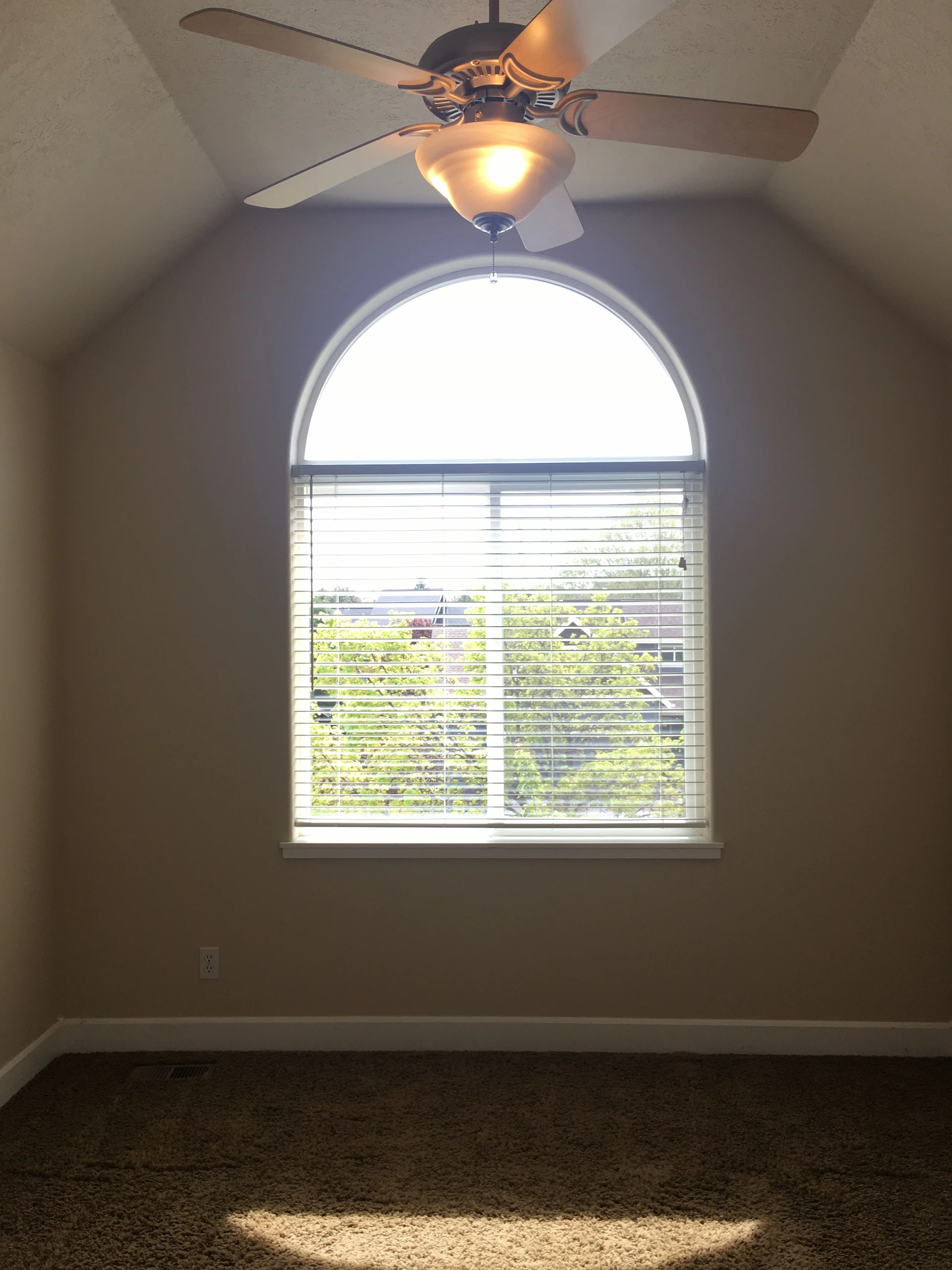 An empty carpeted room has an arched window with blinds and a lit ceiling fan.