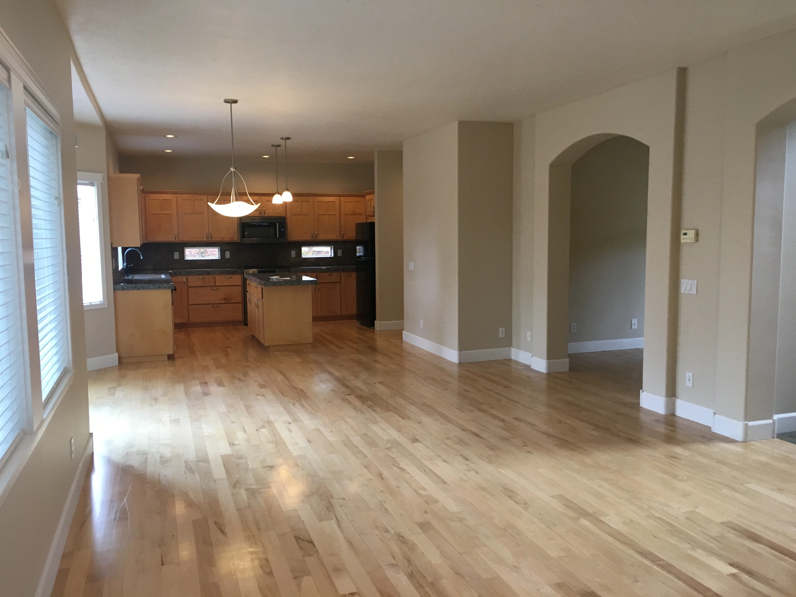 An empty open room with light wood floors, a kitchen with wood cabinets and an island, and an arched doorway.