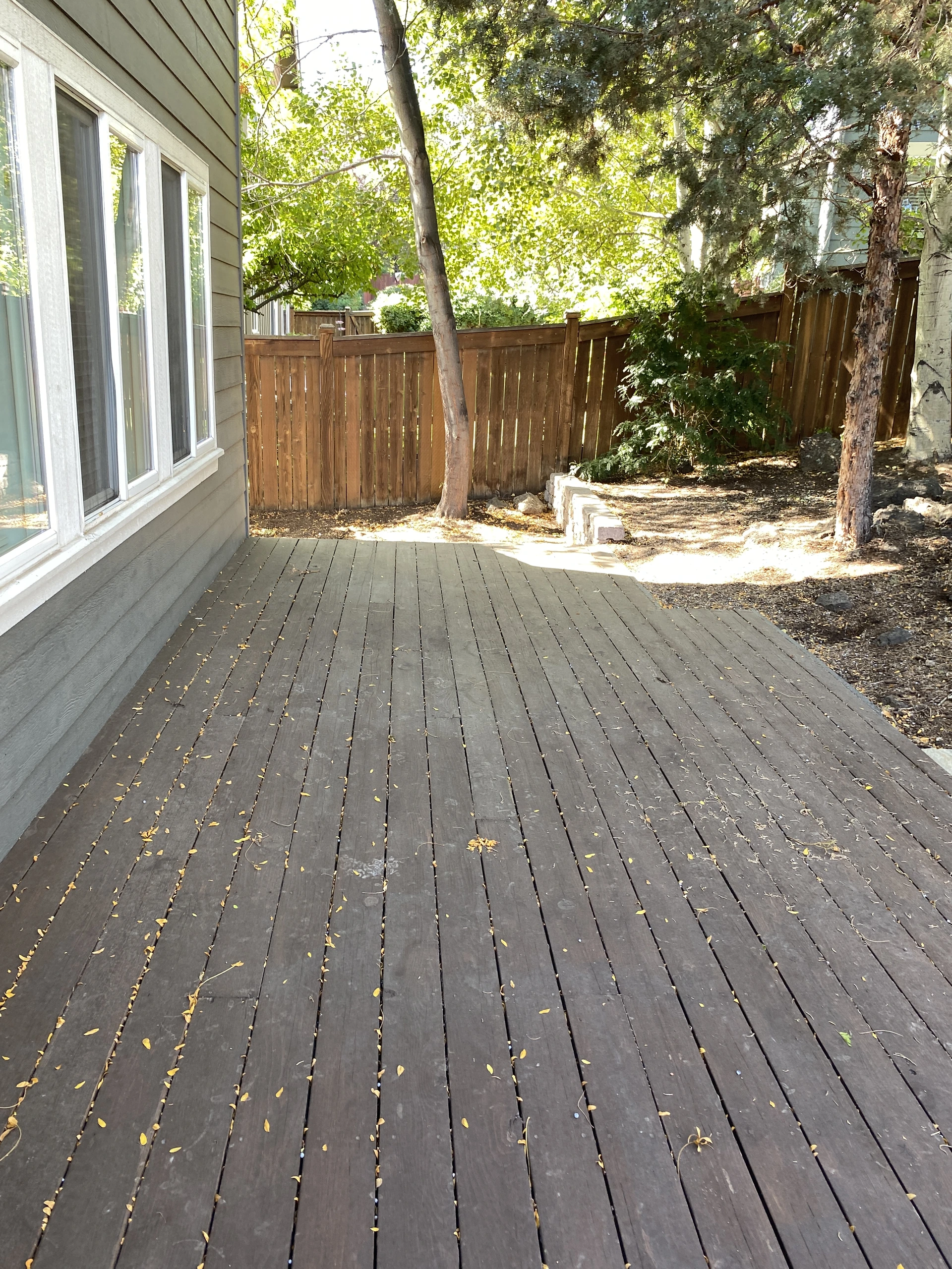 A leaf-strewn wooden deck beside a house, with windows, a wooden fence, and trees around it.