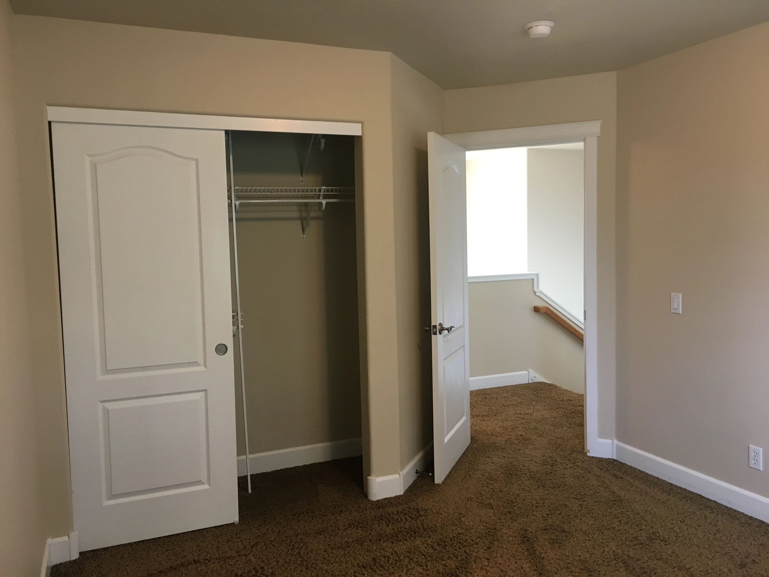 A carpeted room has an open closet with wire shelving and an open doorway to a stair landing.