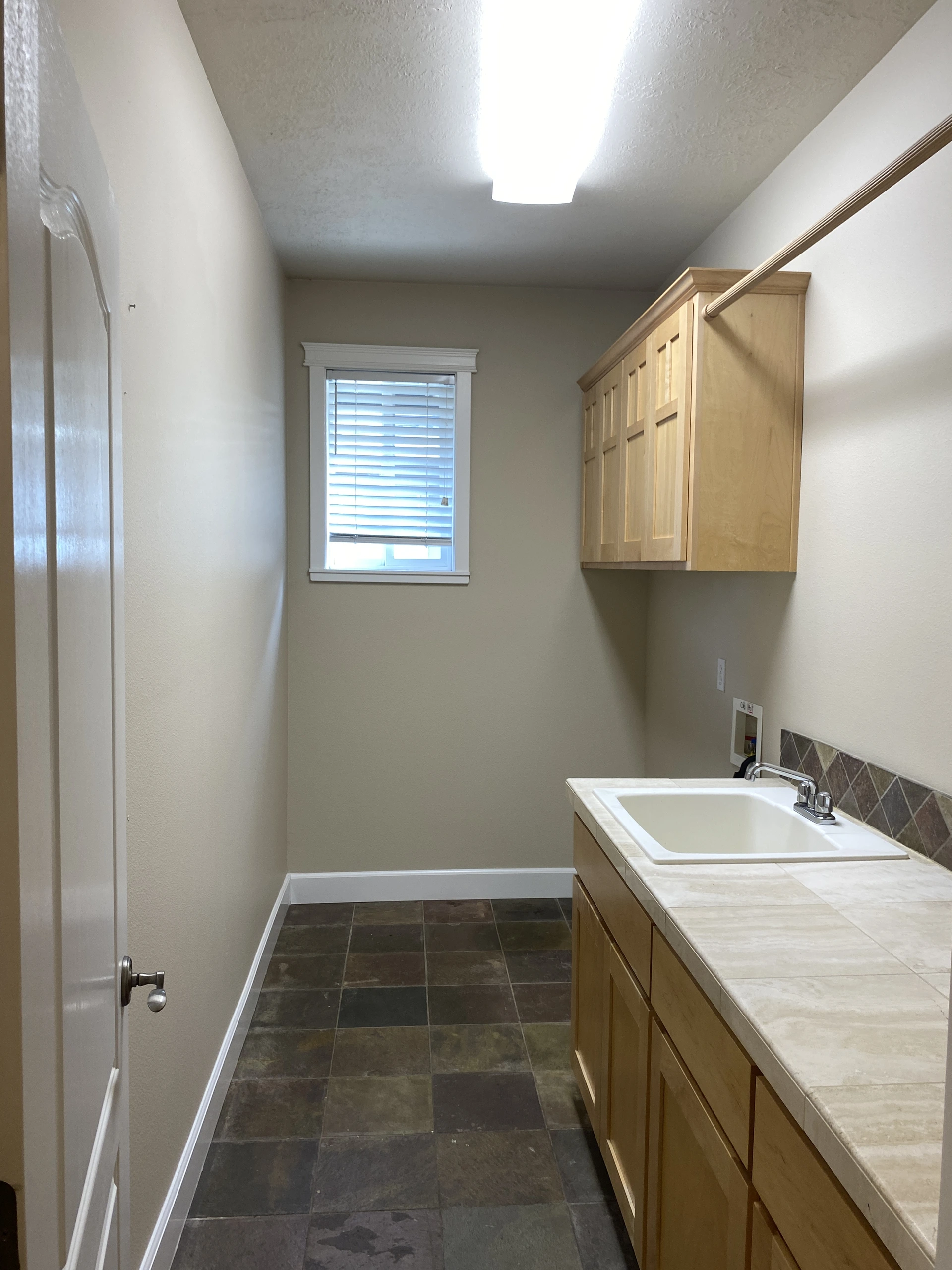 A narrow room has a tiled floor, a window with blinds, and a counter with a sink beneath wooden cabinets.