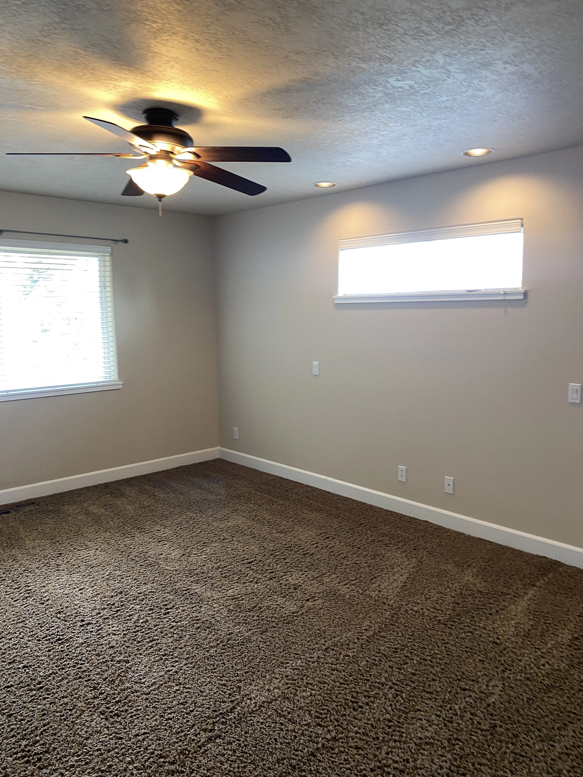 An empty room with brown carpet, two windows with blinds, and a lit ceiling fan beneath recessed lights.