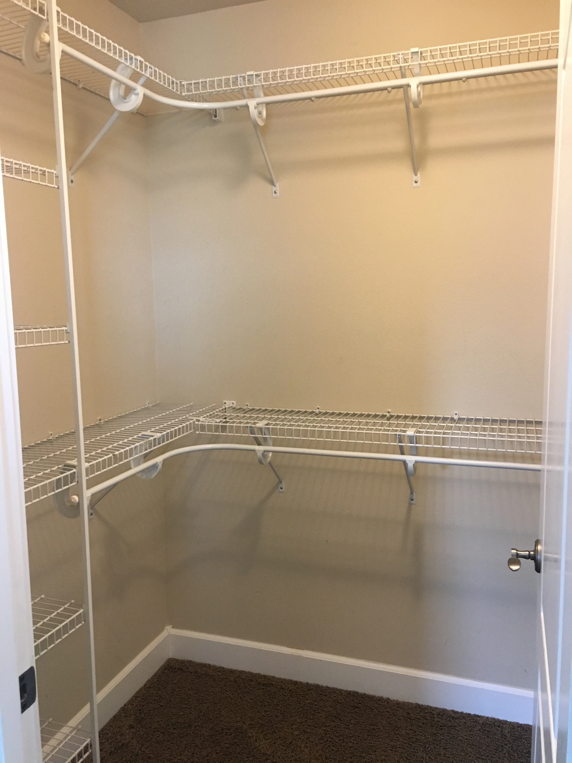 An empty closet has white wire shelves and hanging rods along beige walls, with brown carpet.