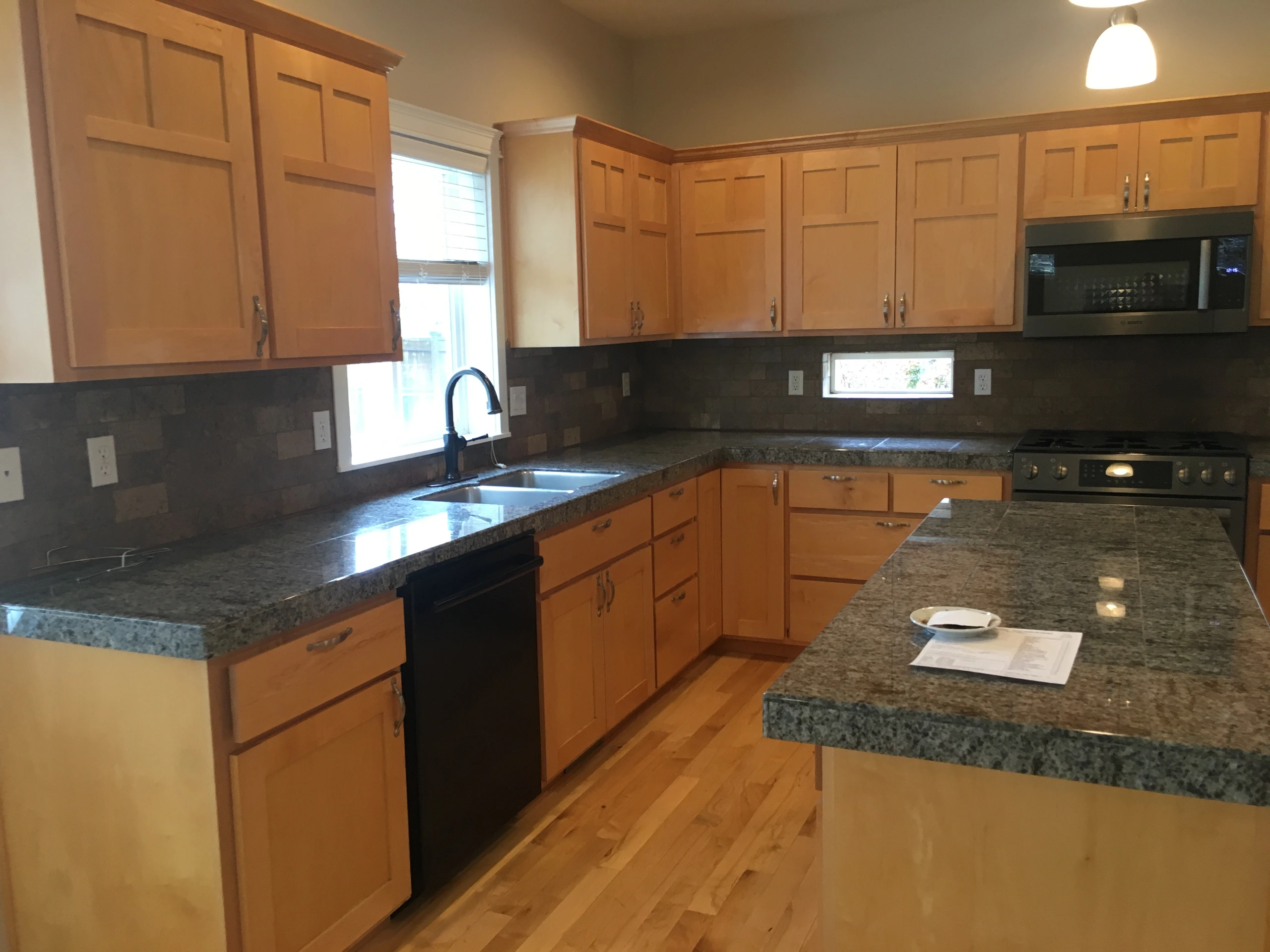 Kitchen with honey-colored cabinets, dark stone counters, a sink beneath a window, and a central island.