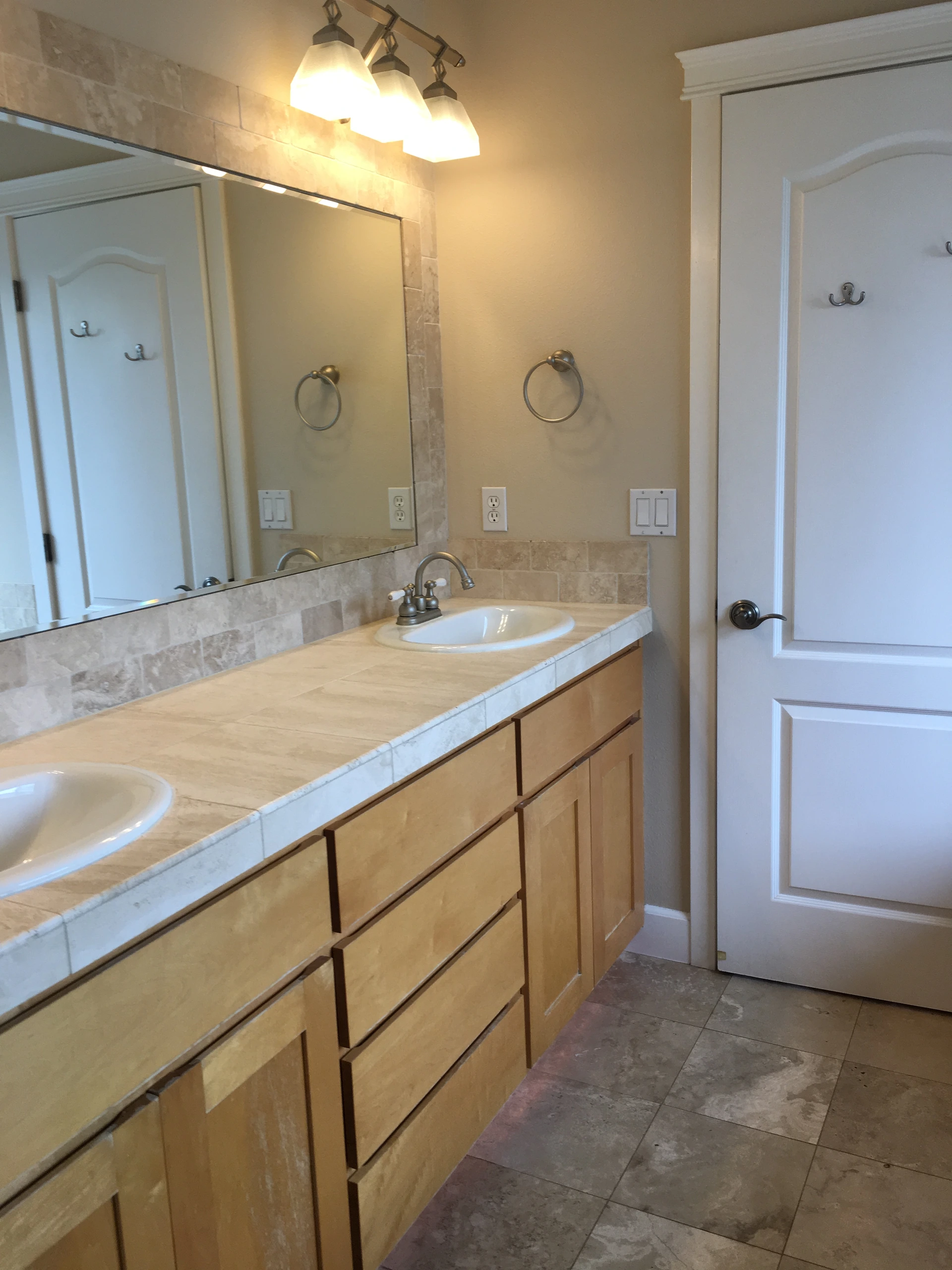 A tiled bathroom vanity with two sinks, a large mirror, wooden drawers, three lights, and a white door.