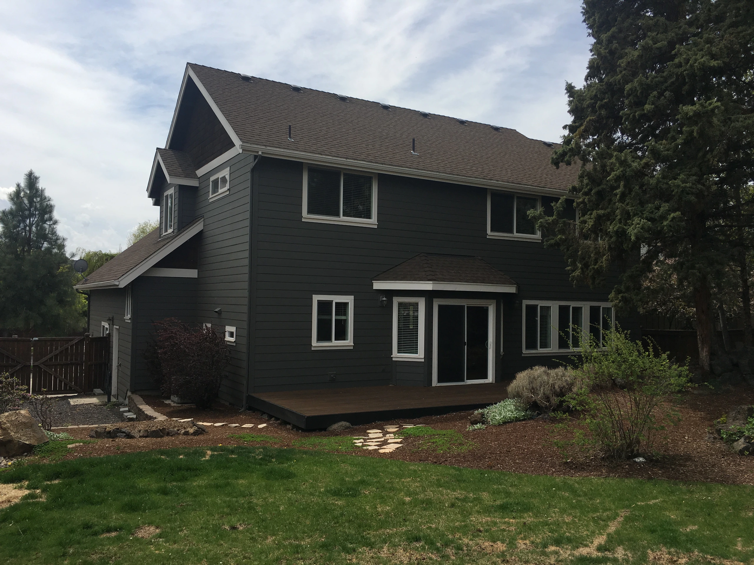 A dark-sided two-story house has white-trimmed windows, a sliding glass door, and a wooden deck facing a landscaped yard.
