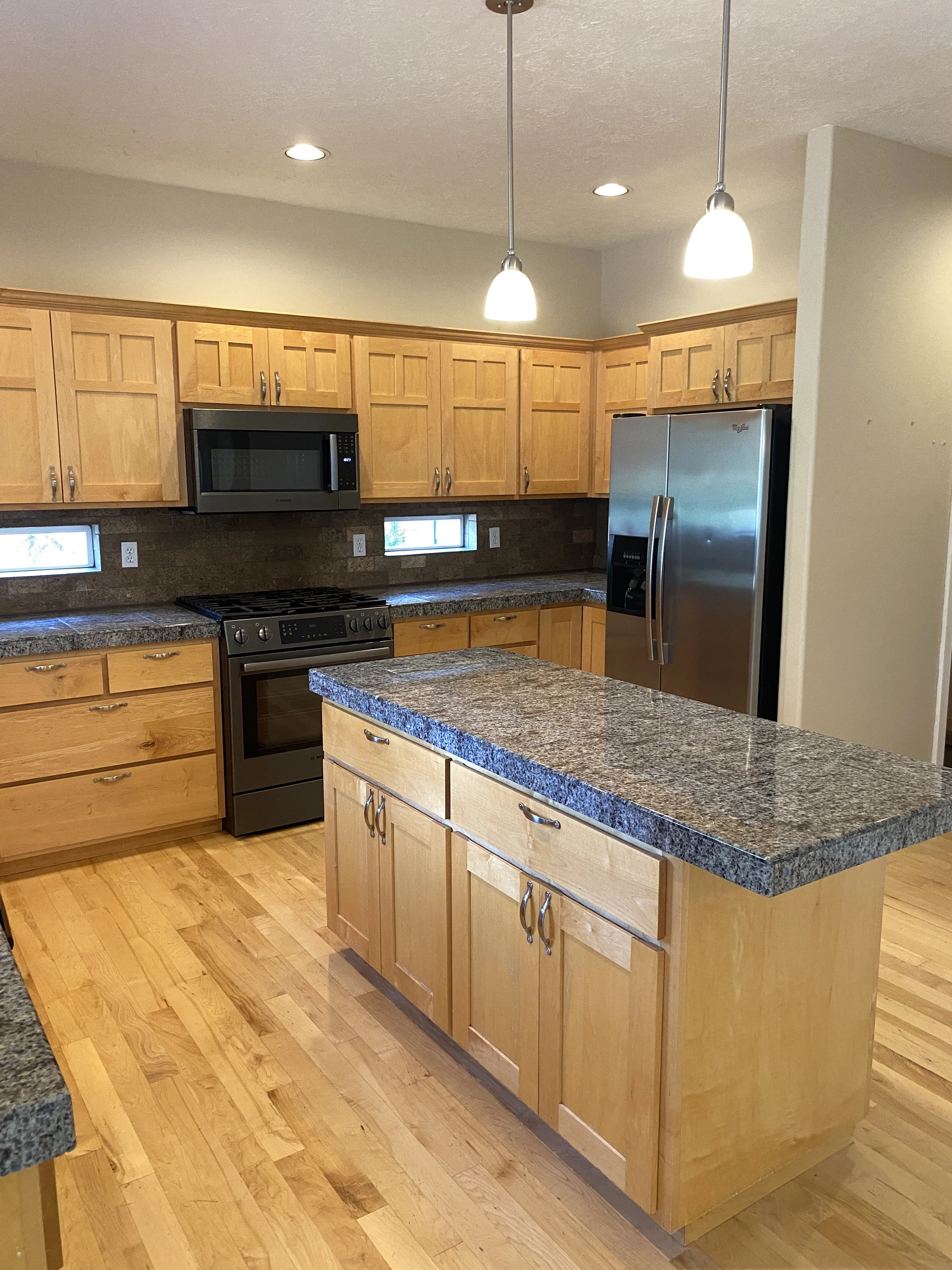 A kitchen with light wood cabinets, a stone-topped island, a stove, a microwave, and a stainless steel refrigerator.