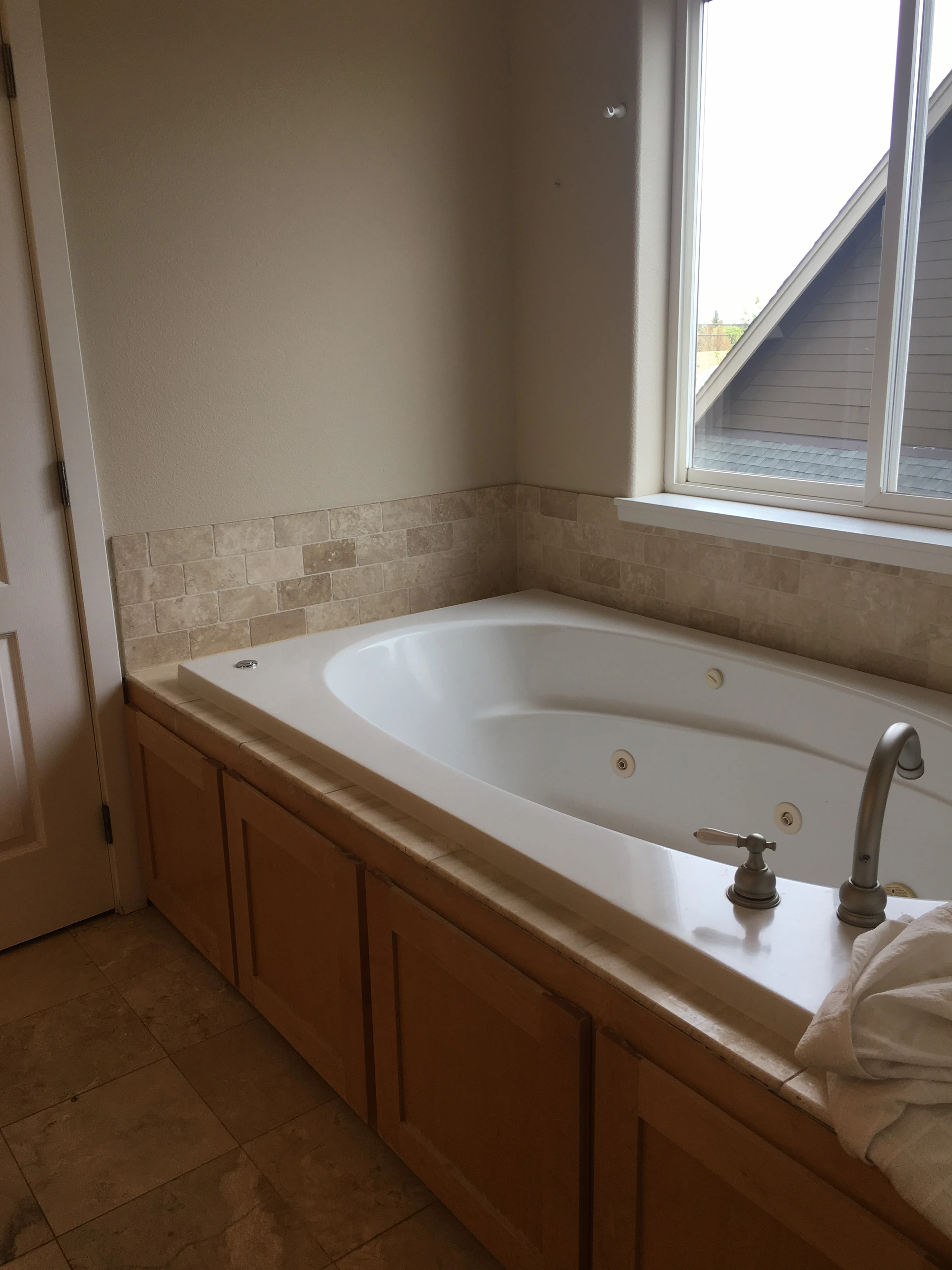 A white jetted bathtub with a tall faucet sits beneath a large window, bordered by beige tile and wood-paneled cabinetry.