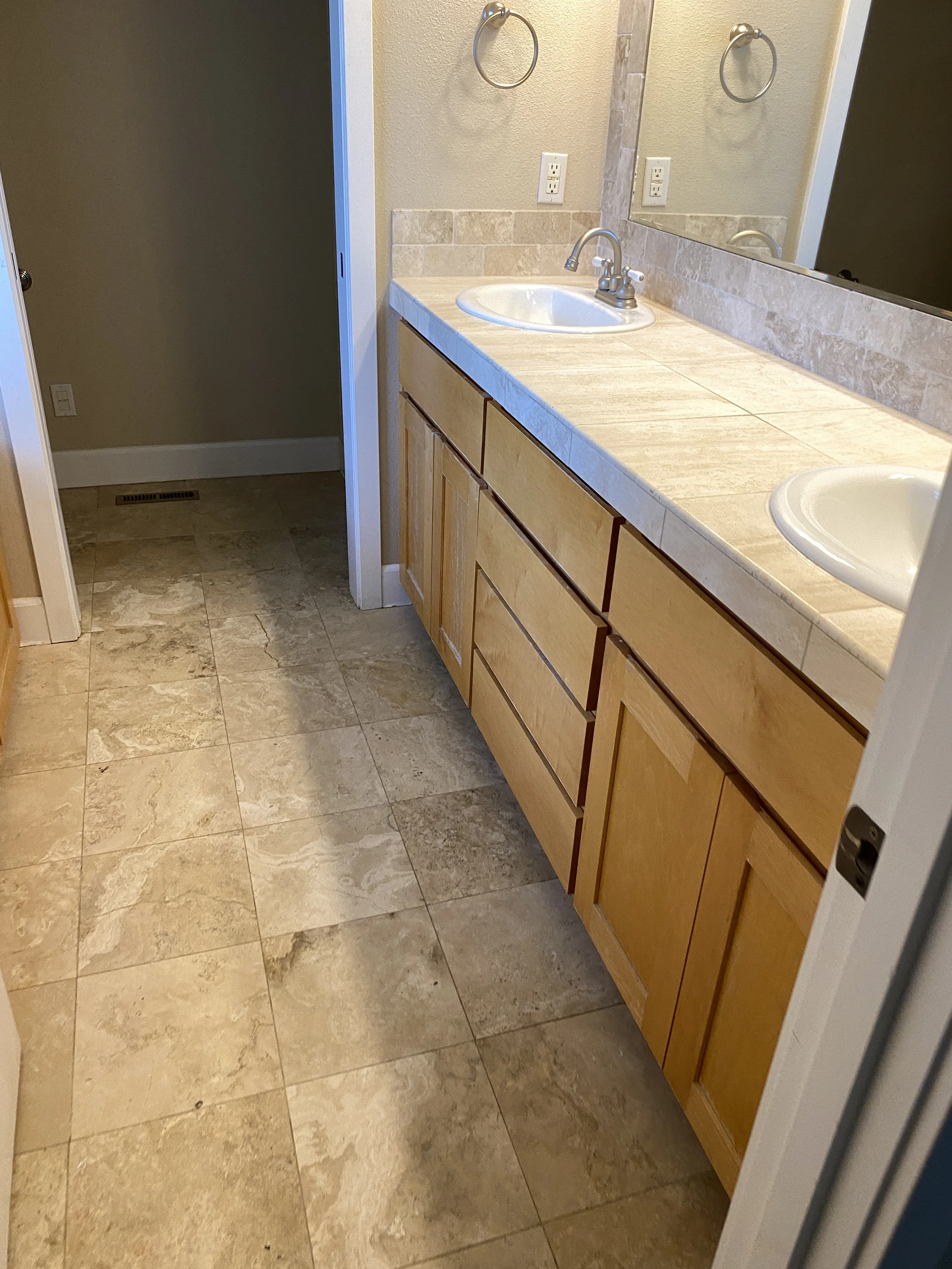 A tiled bathroom vanity has a long wooden cabinet, two white sinks, a large mirror, and metal towel rings.