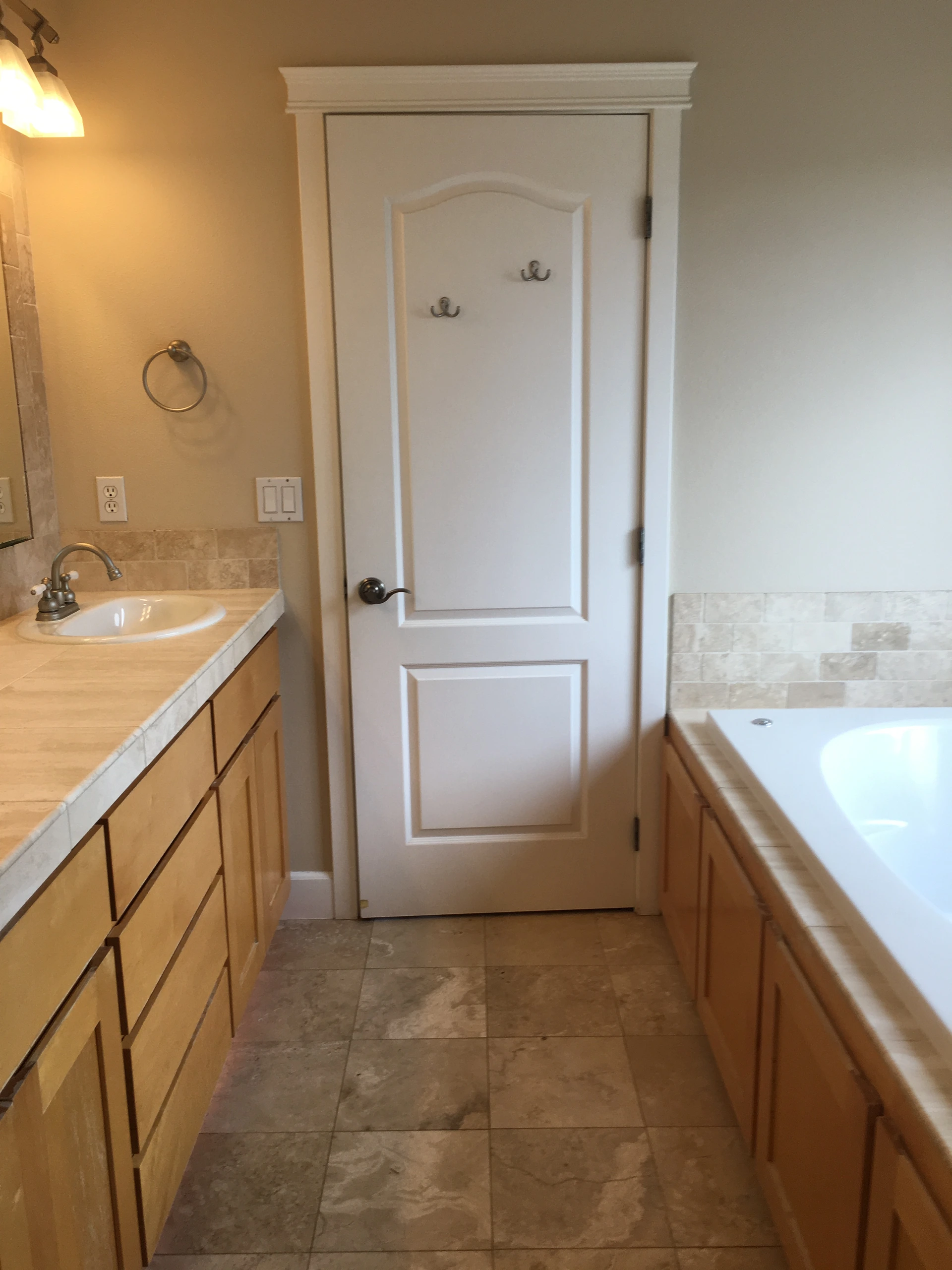 A bathroom with a white paneled door between a sink vanity and a bathtub, with tile flooring and wall accents.