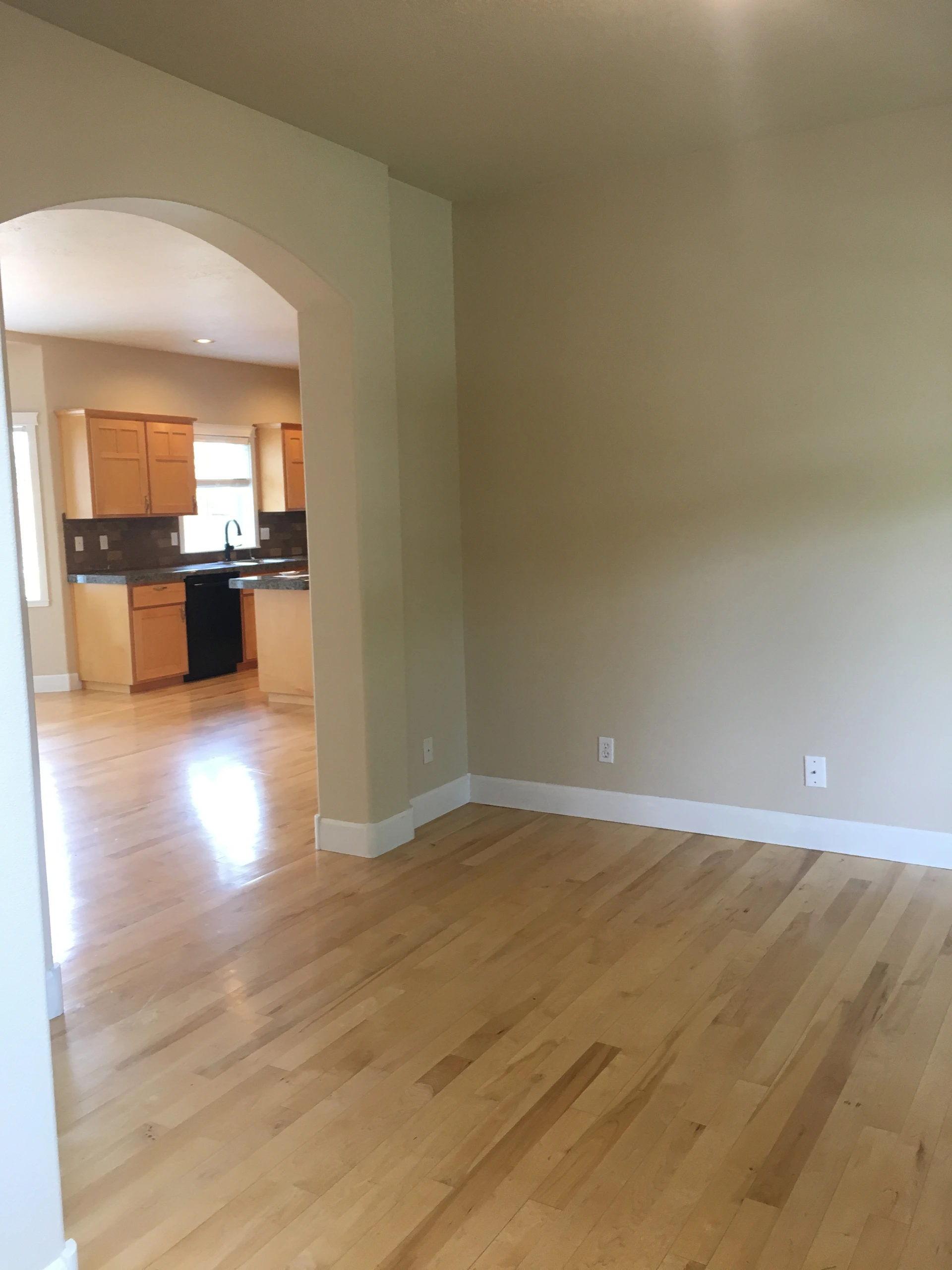 An empty room with light wood flooring and an arched opening into a kitchen with wooden cabinets and a window.
