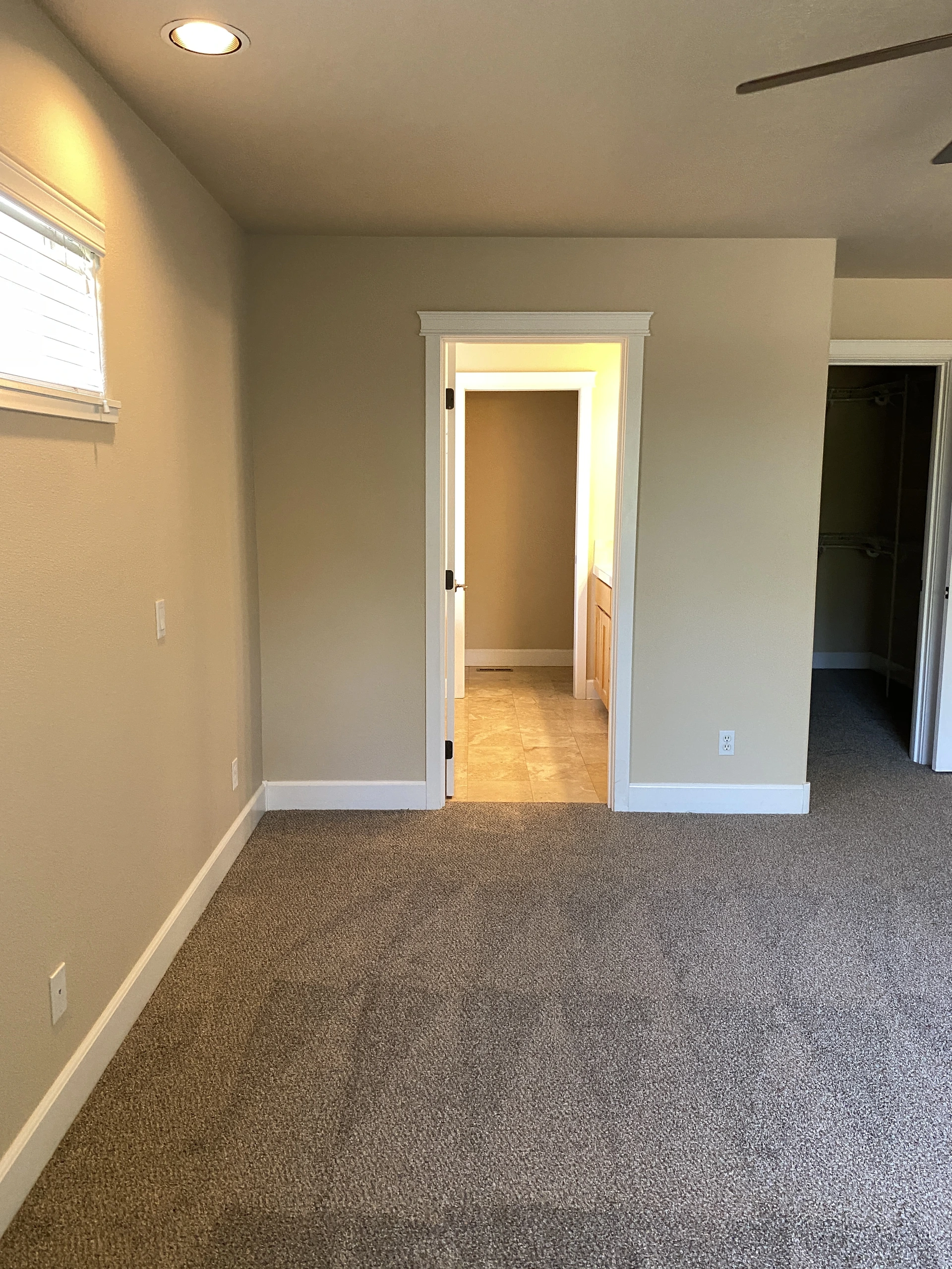 An empty carpeted room has beige walls, a window with blinds, and two open doorways.