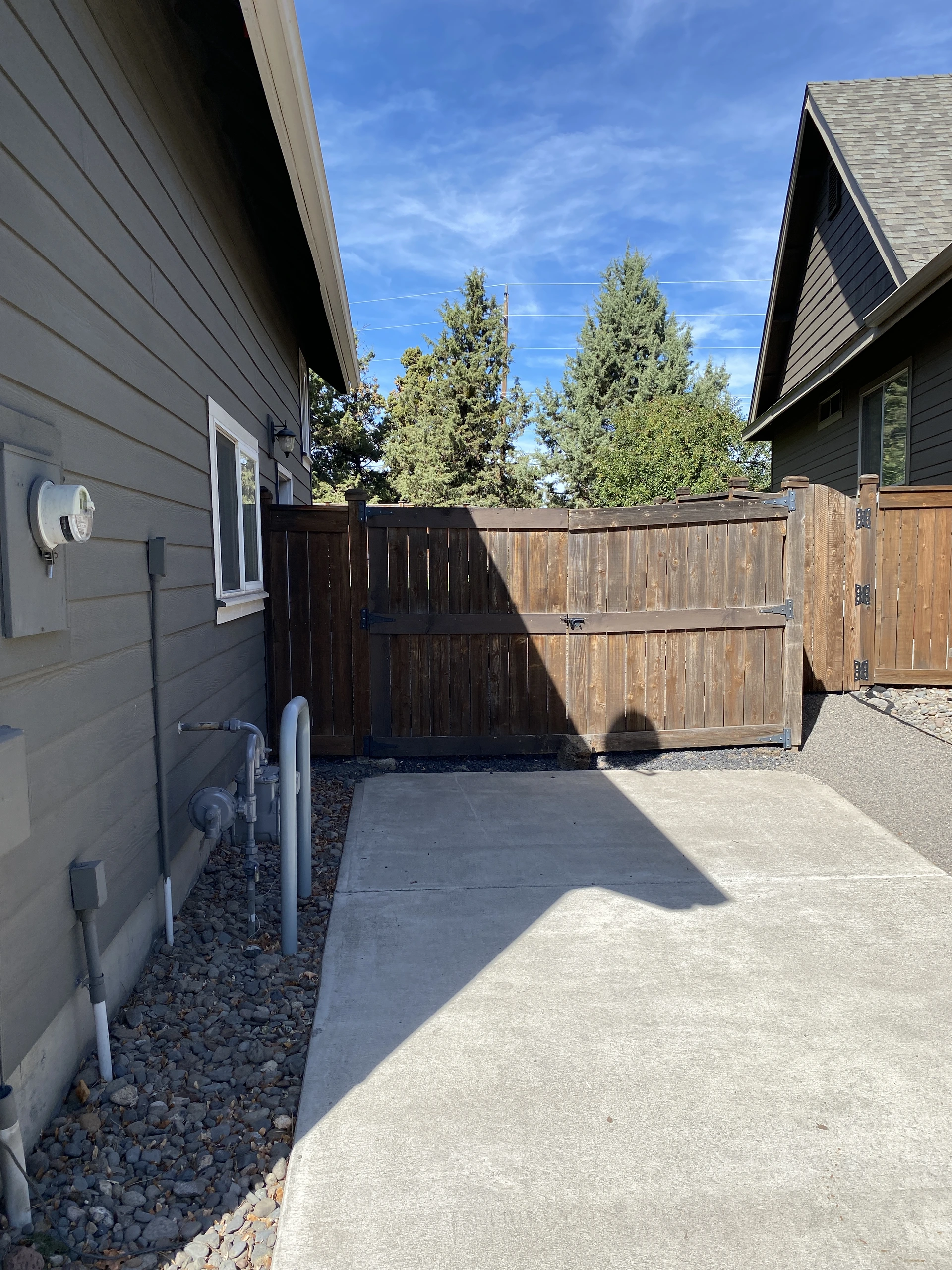 A concrete walkway between two houses ends at a tall wooden gate and fence, with trees visible beyond.