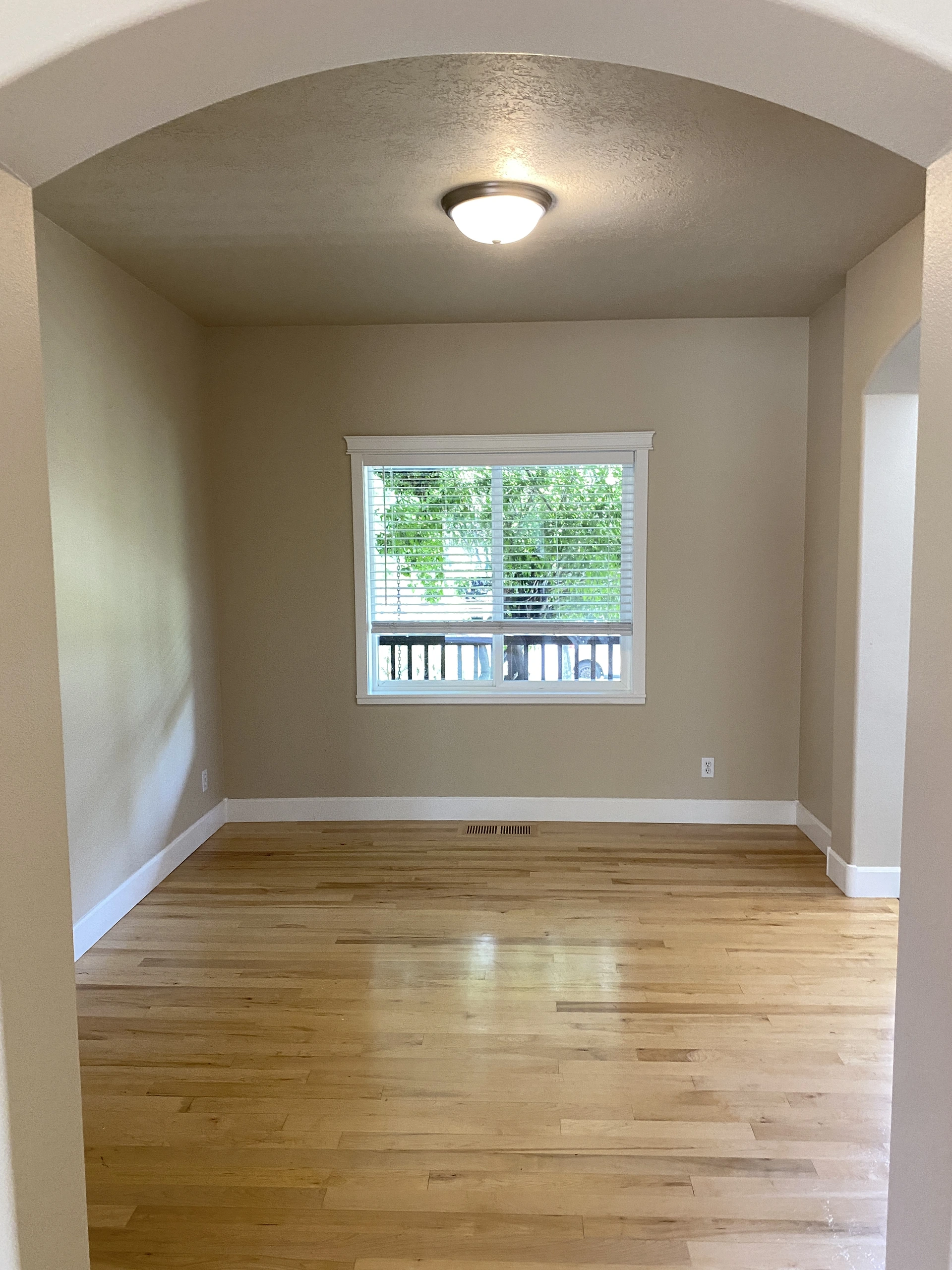 An empty room with light wood flooring, beige walls, a window with blinds, and a ceiling light.