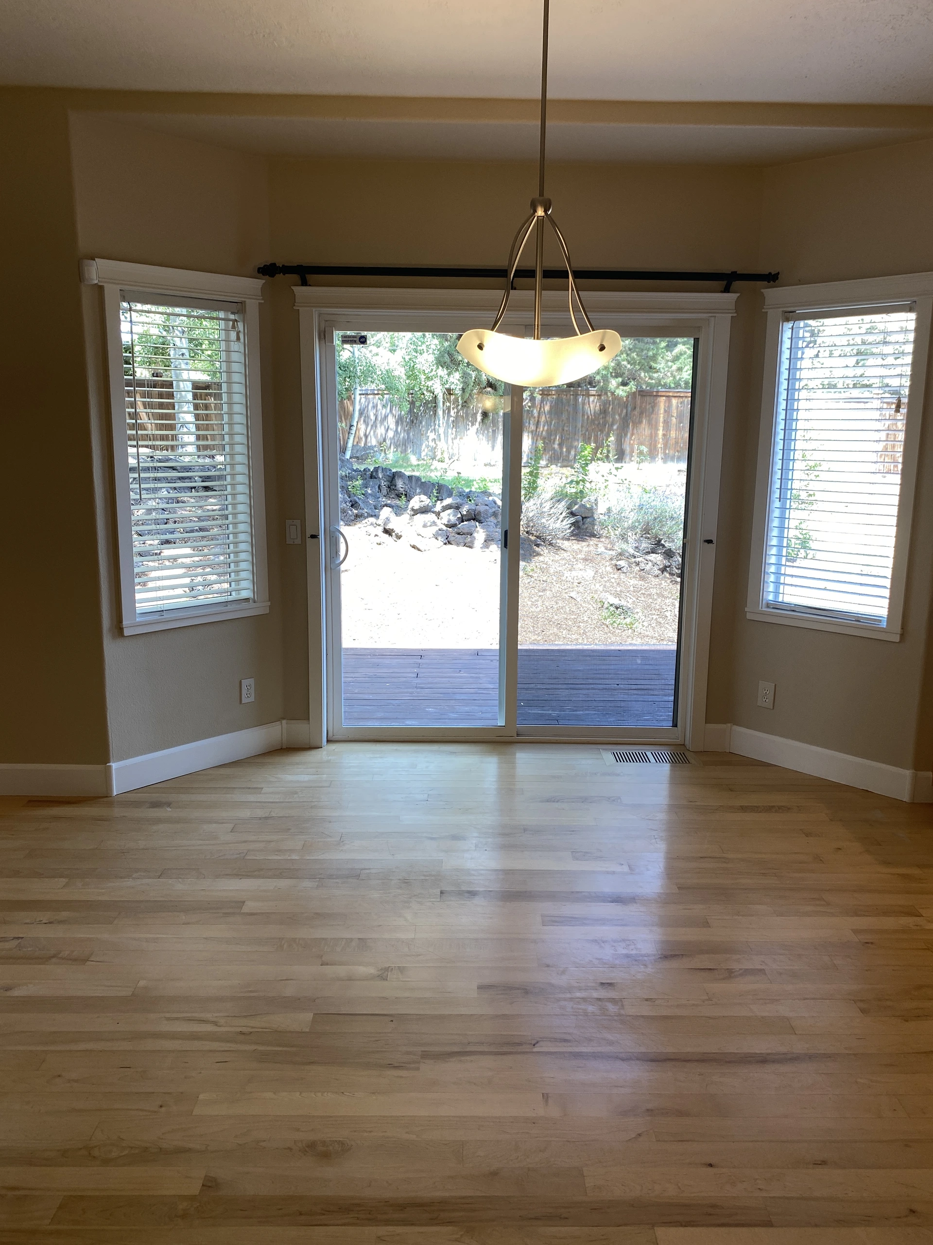 An empty room with light wood flooring, a central sliding glass door, two side windows with blinds, and a hanging light.