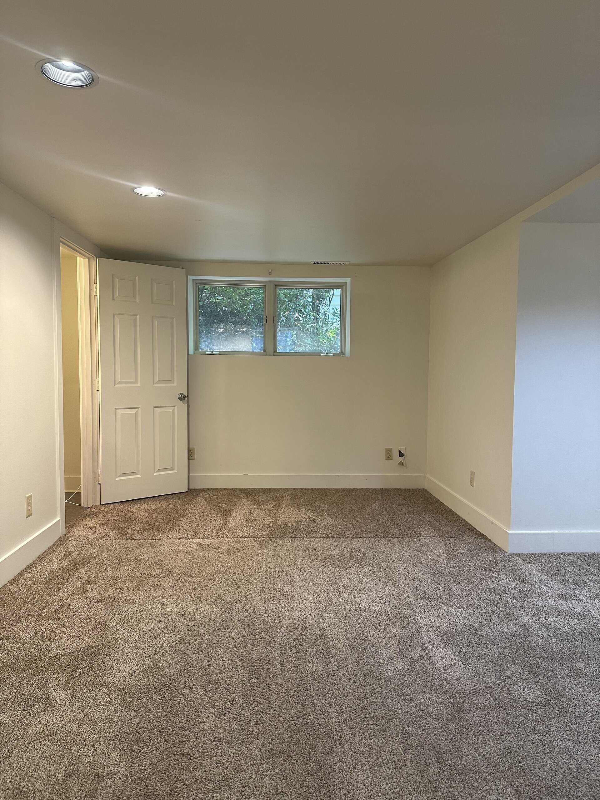An empty room with brown carpet, beige walls, recessed ceiling lights, a small window, and an open door.