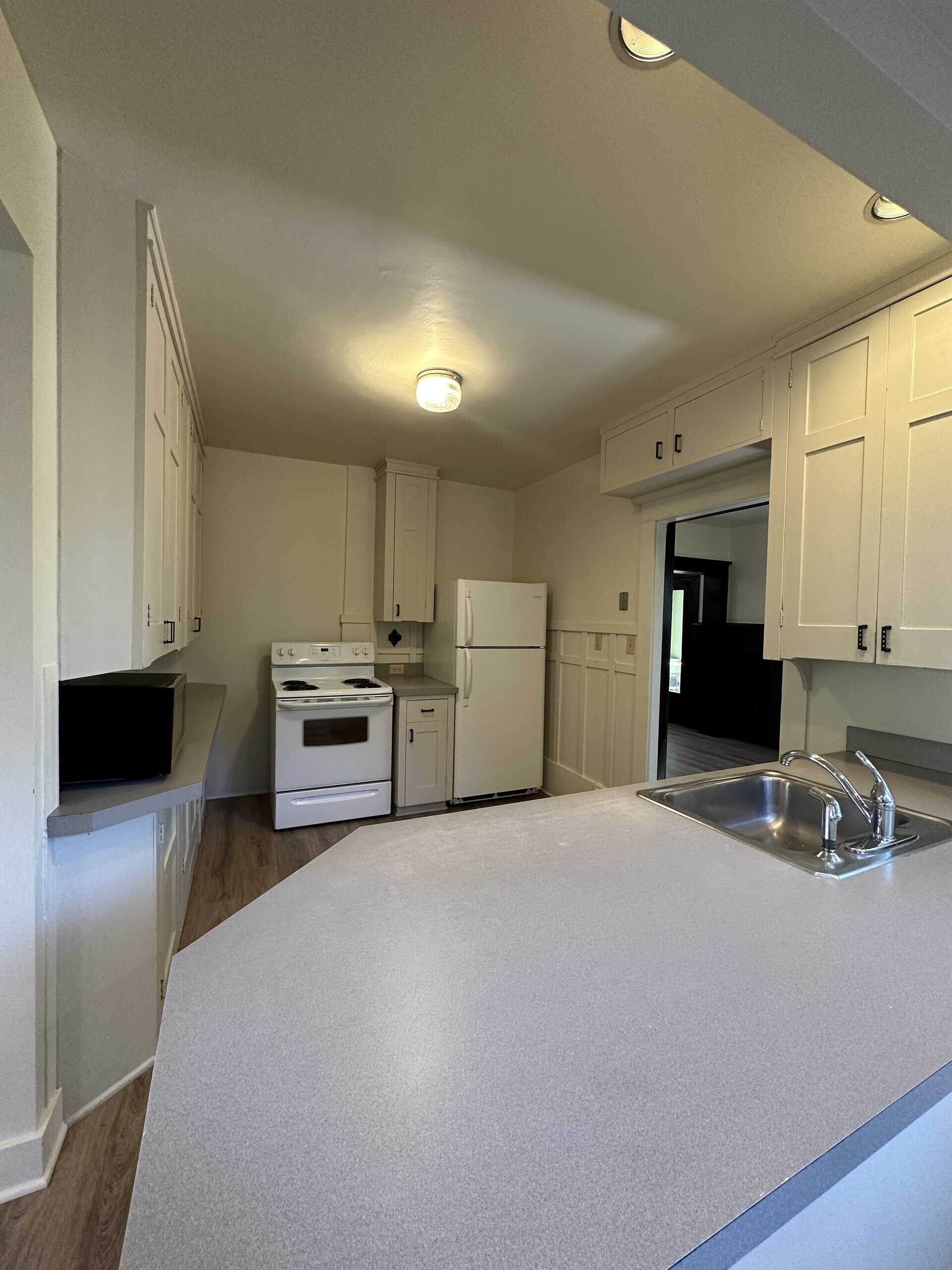 A kitchen with white cabinets, a stove and refrigerator, and a sink set into a large foreground counter.