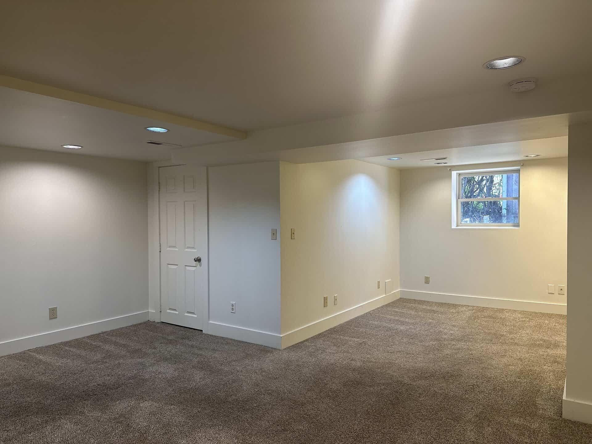 An empty carpeted room has cream walls, a white door, a small window, and recessed ceiling lights.