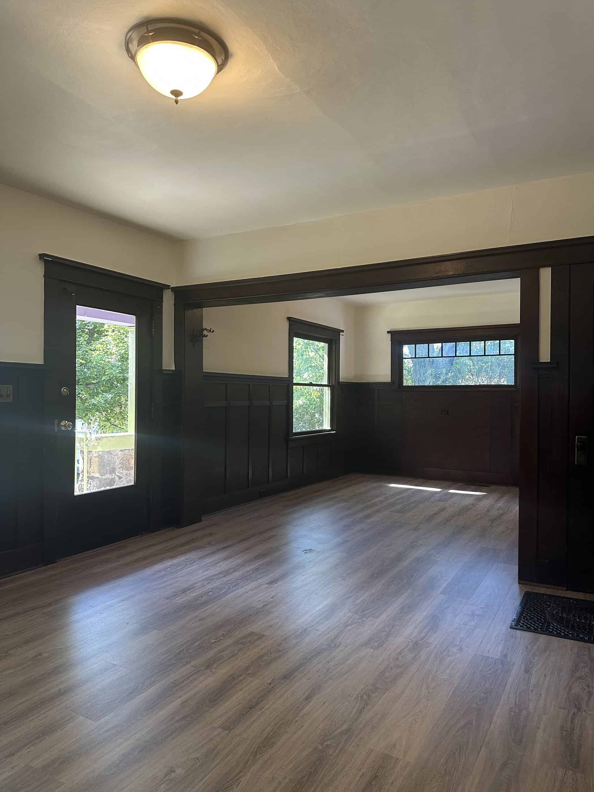 An empty room has dark wood wall paneling, several windows, a ceiling light, and gray-brown plank flooring.