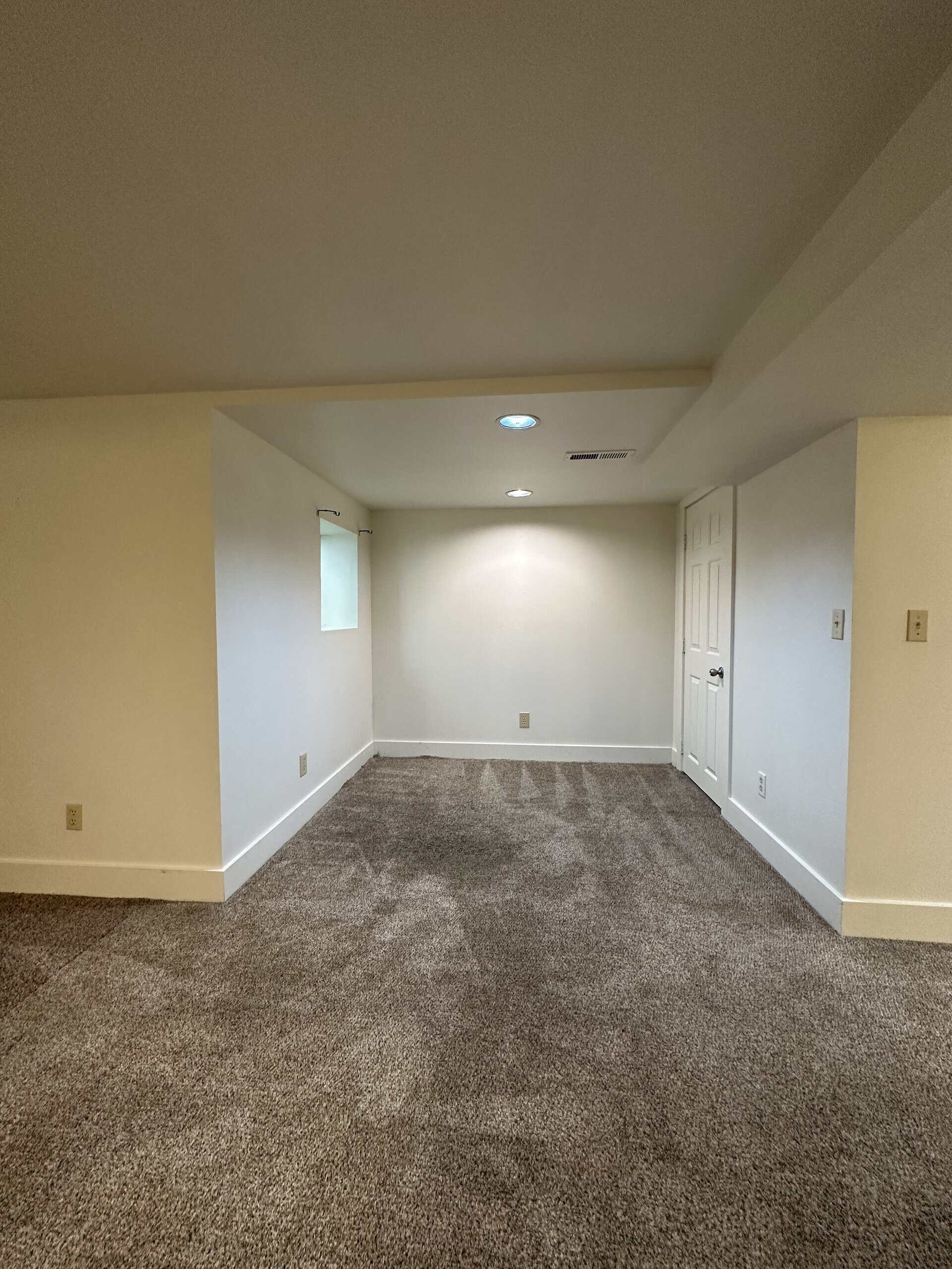 An empty carpeted room with pale walls, recessed ceiling lights, a small window, and a closed door.