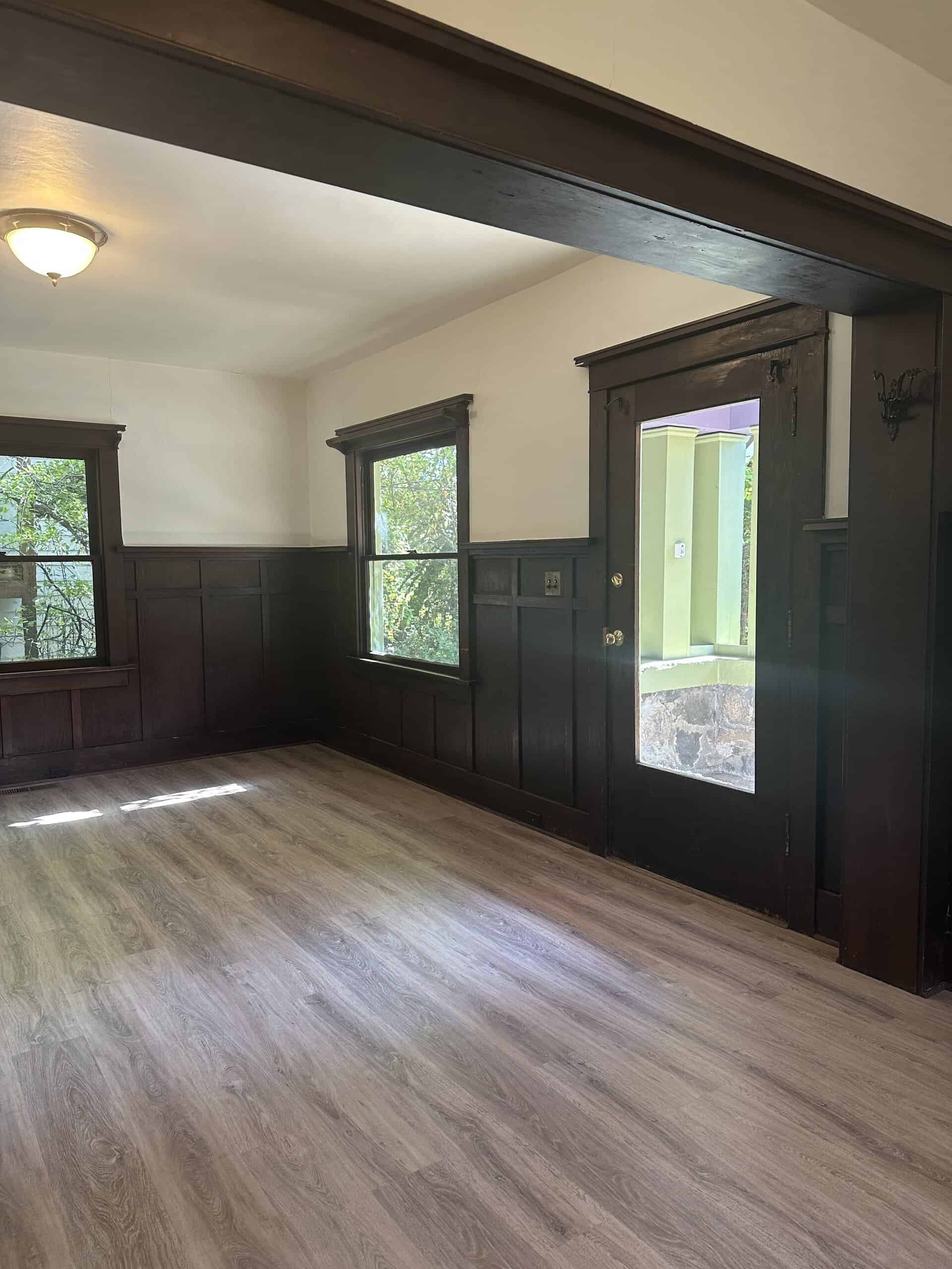 An empty room has dark wood wall paneling, two windows, an open doorway, light wood-patterned flooring, and a ceiling light.