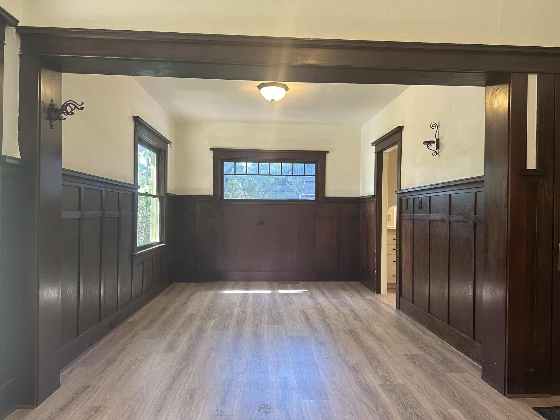 An empty room has dark wood wall paneling, light wood flooring, two windows, and an open doorway.
