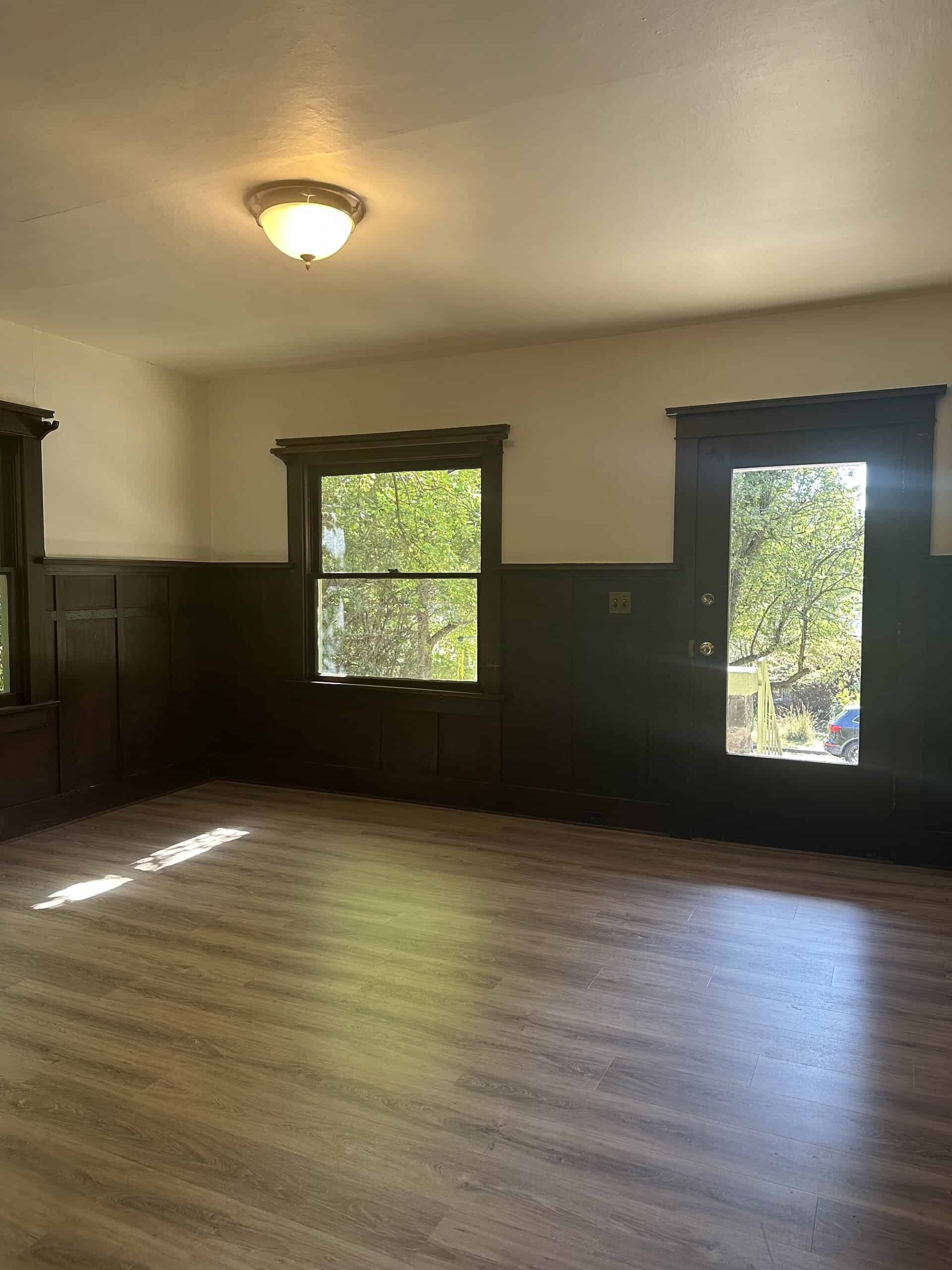 An empty room has dark wood wall paneling, a window, a glass-paneled door, wood-look flooring, and a lit ceiling fixture.