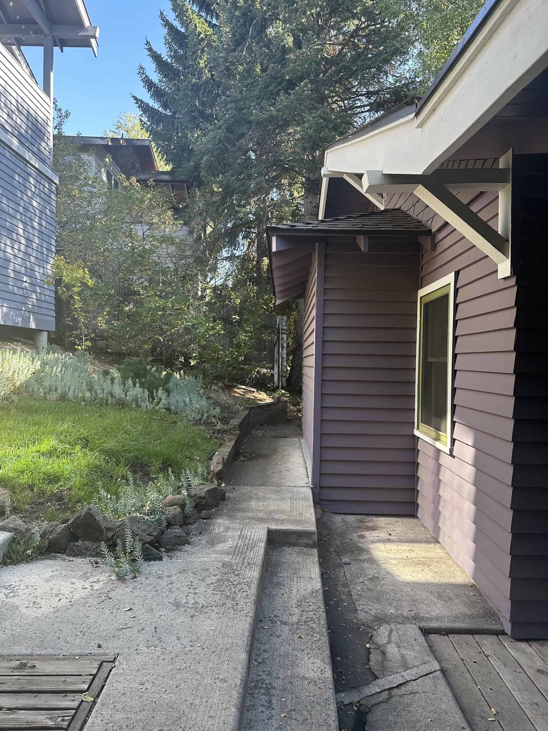 A concrete walkway runs beside a mauve-sided building with a window, bordered by rocks, plants, and trees.