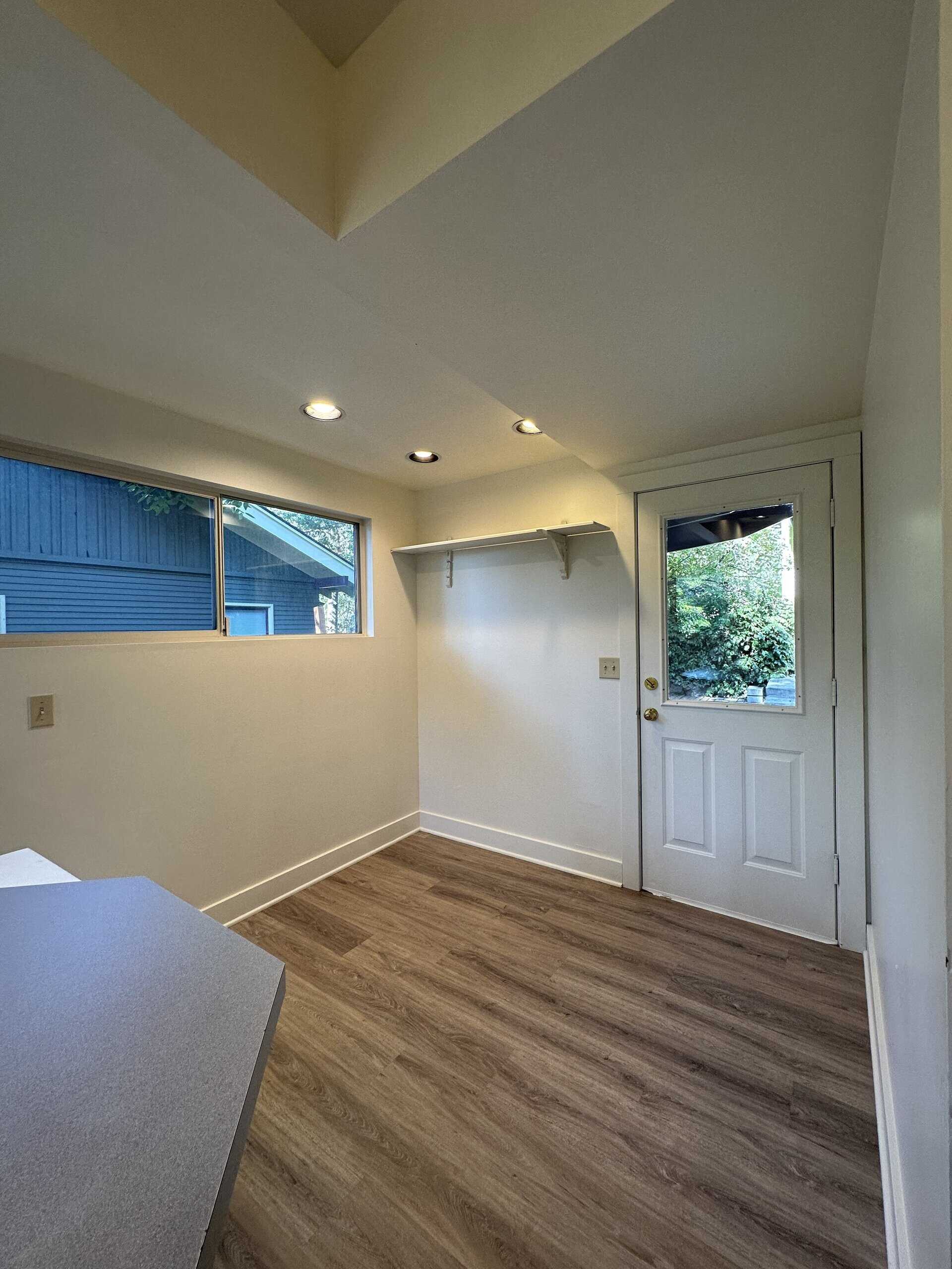 A small room with wood-patterned flooring, a high horizontal window, a wall shelf, and a white door with a window.