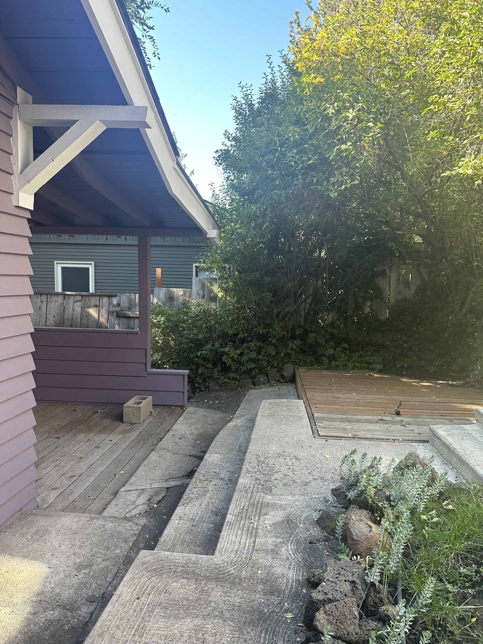 A covered porch beside a concrete path and wooden deck, with leafy trees along the right side.
