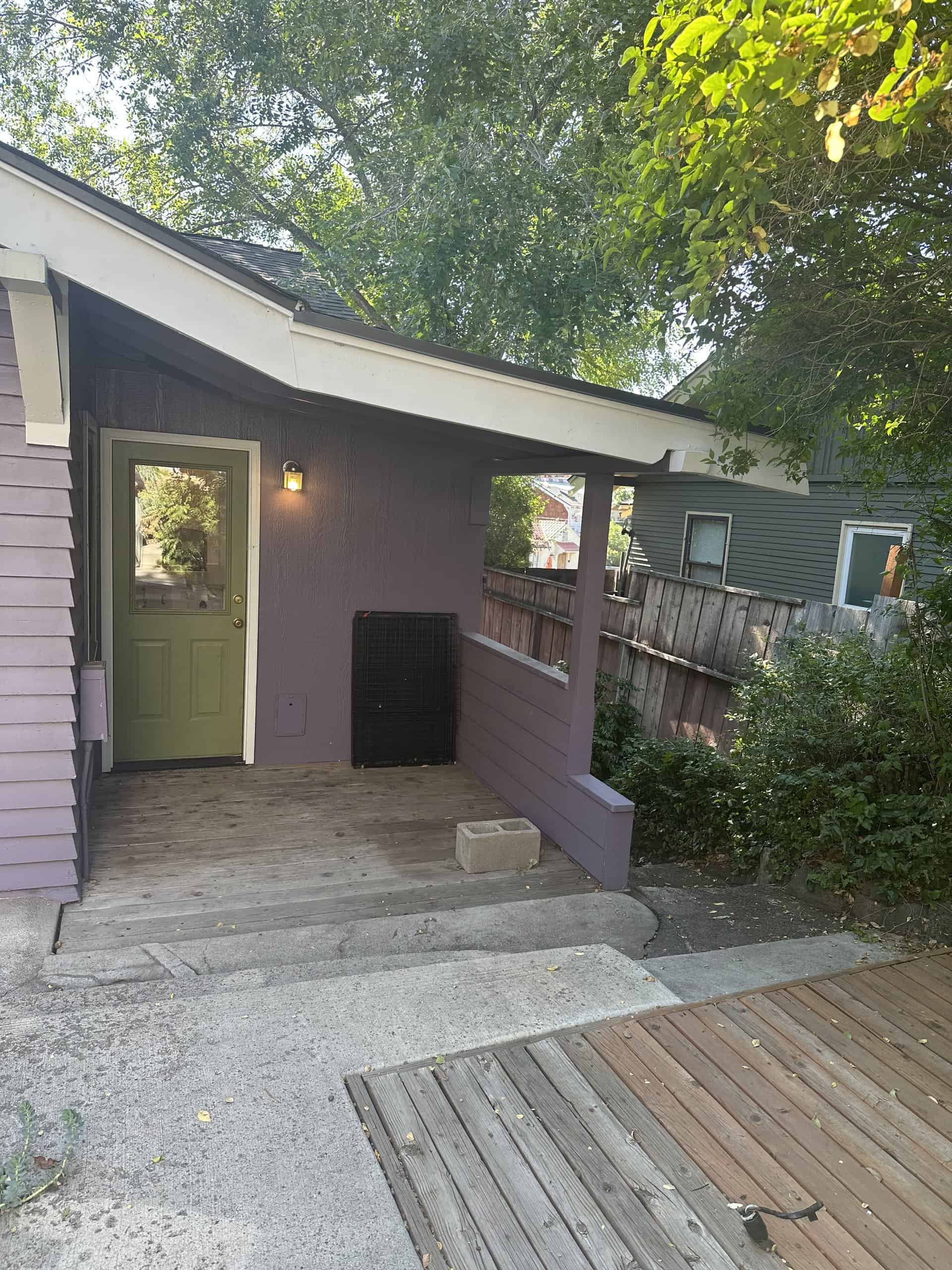 A covered porch has a green door, a wooden deck, and leafy trees overhead.