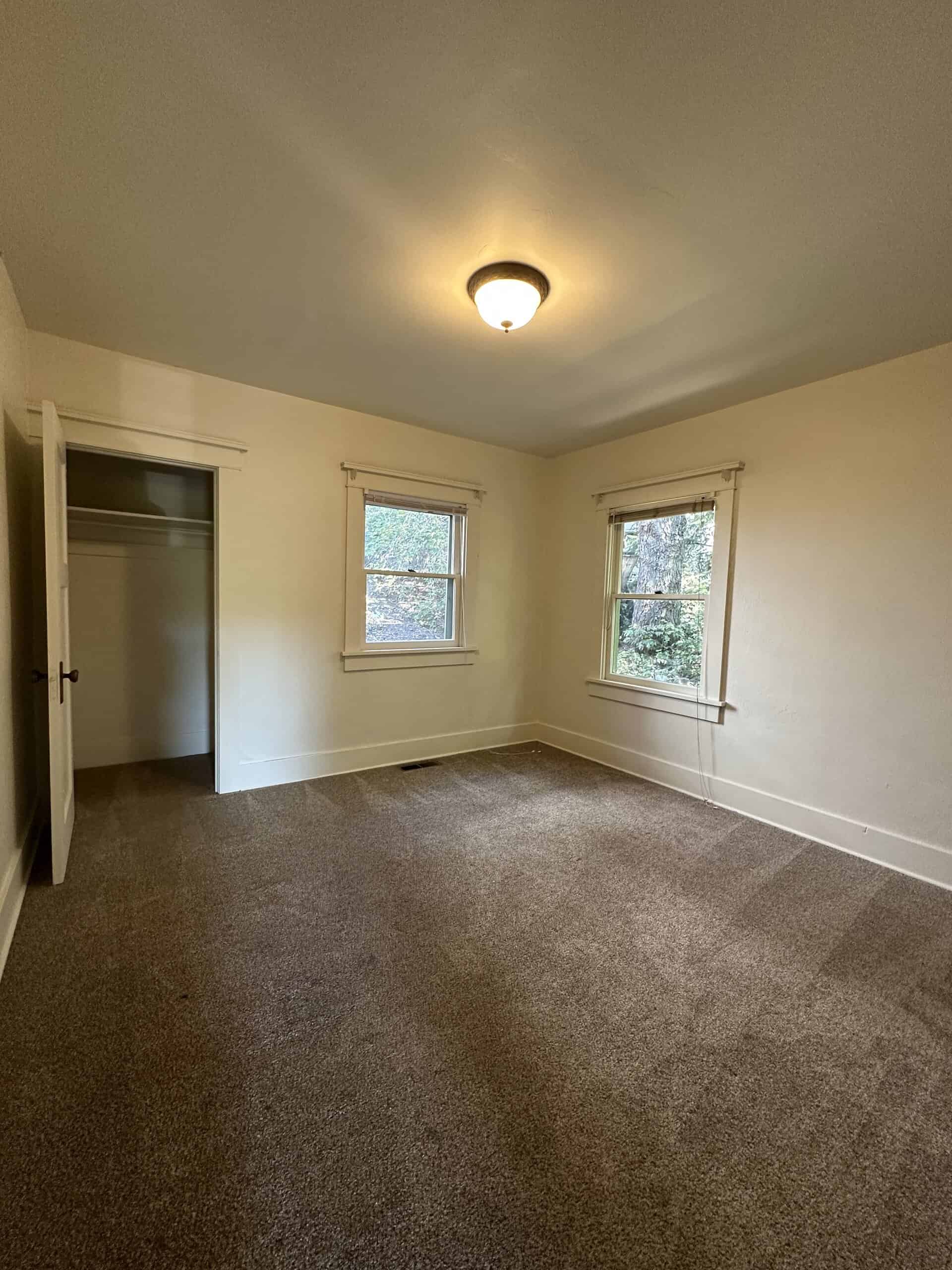 An empty room with brown carpet, two windows, an open closet, and a lit ceiling fixture.