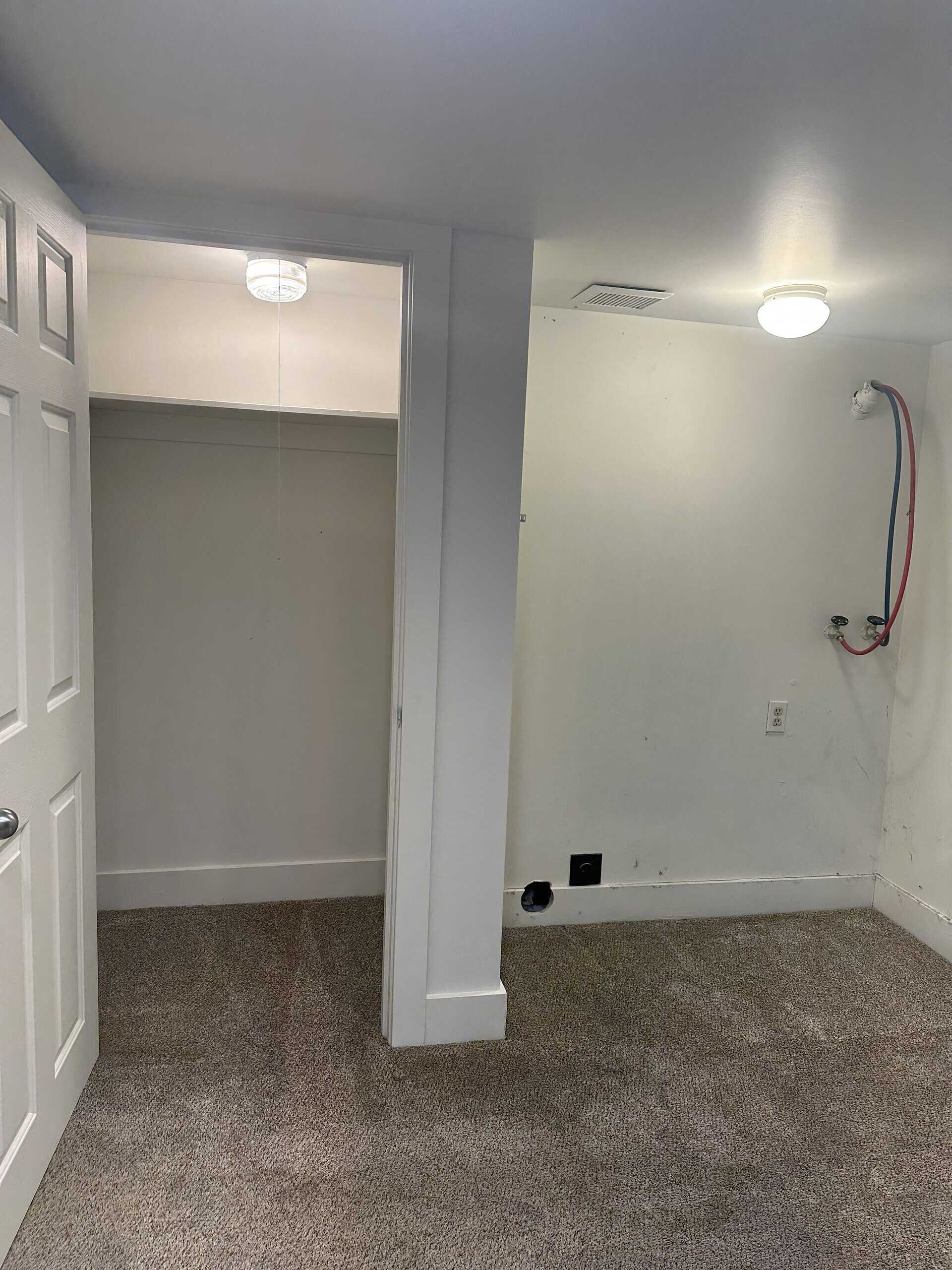 A carpeted room has an open closet, two ceiling lights, and red and blue hoses connected to wall fittings.
