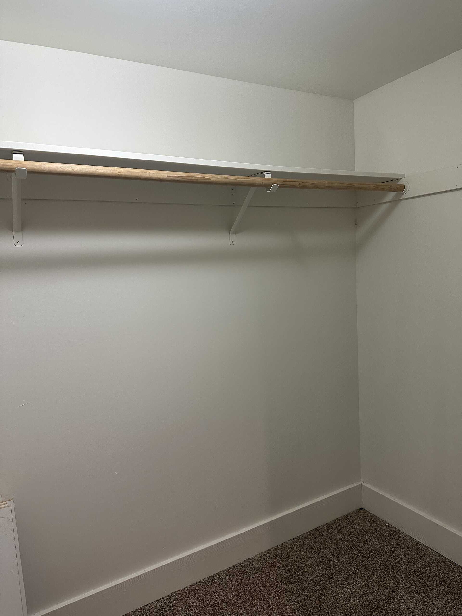 A wooden hanging rod runs beneath a white shelf in a carpeted corner with plain light-colored walls.