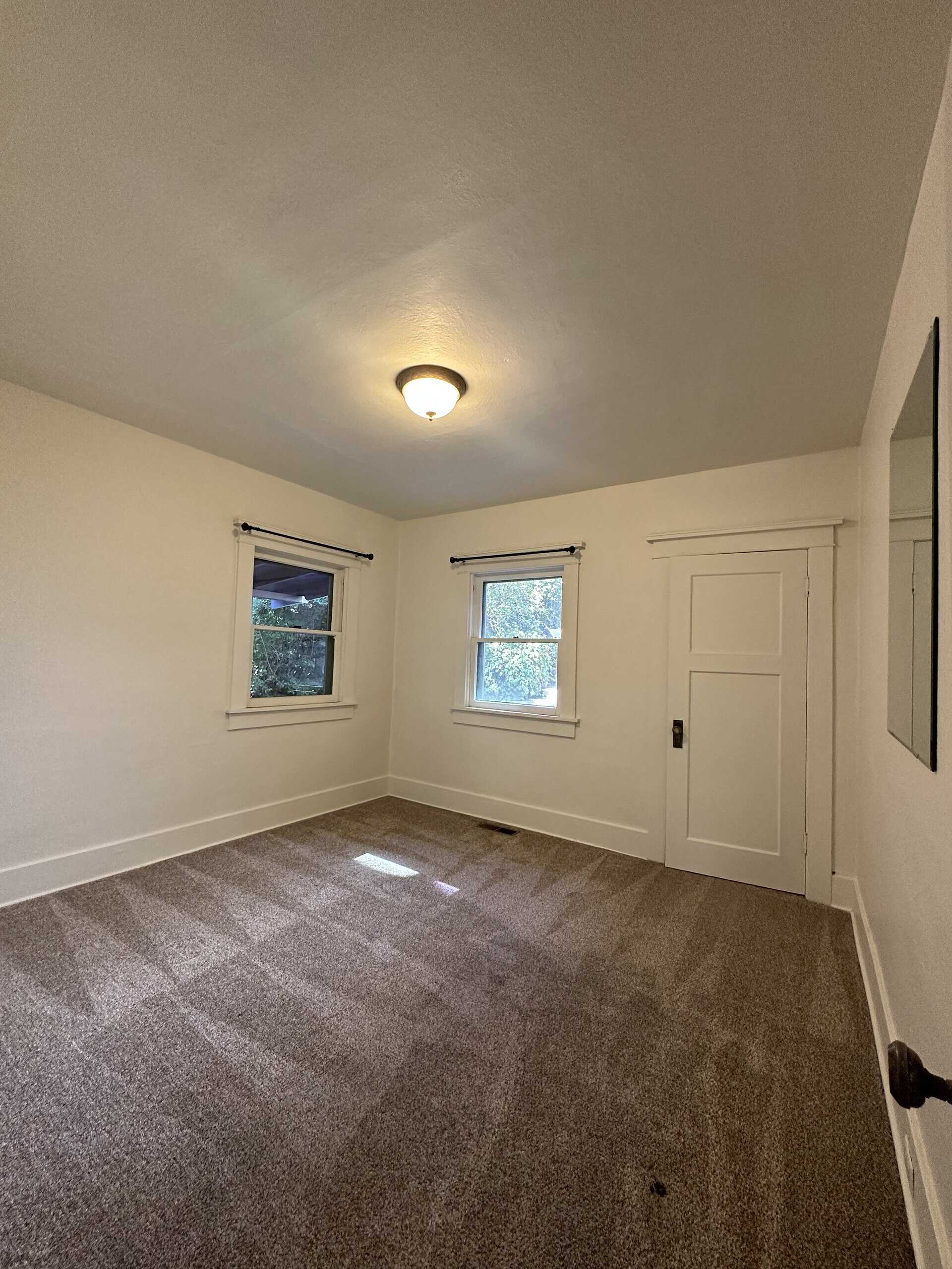 An empty room with brown carpet, two windows, a paneled door, and a lit ceiling fixture.
