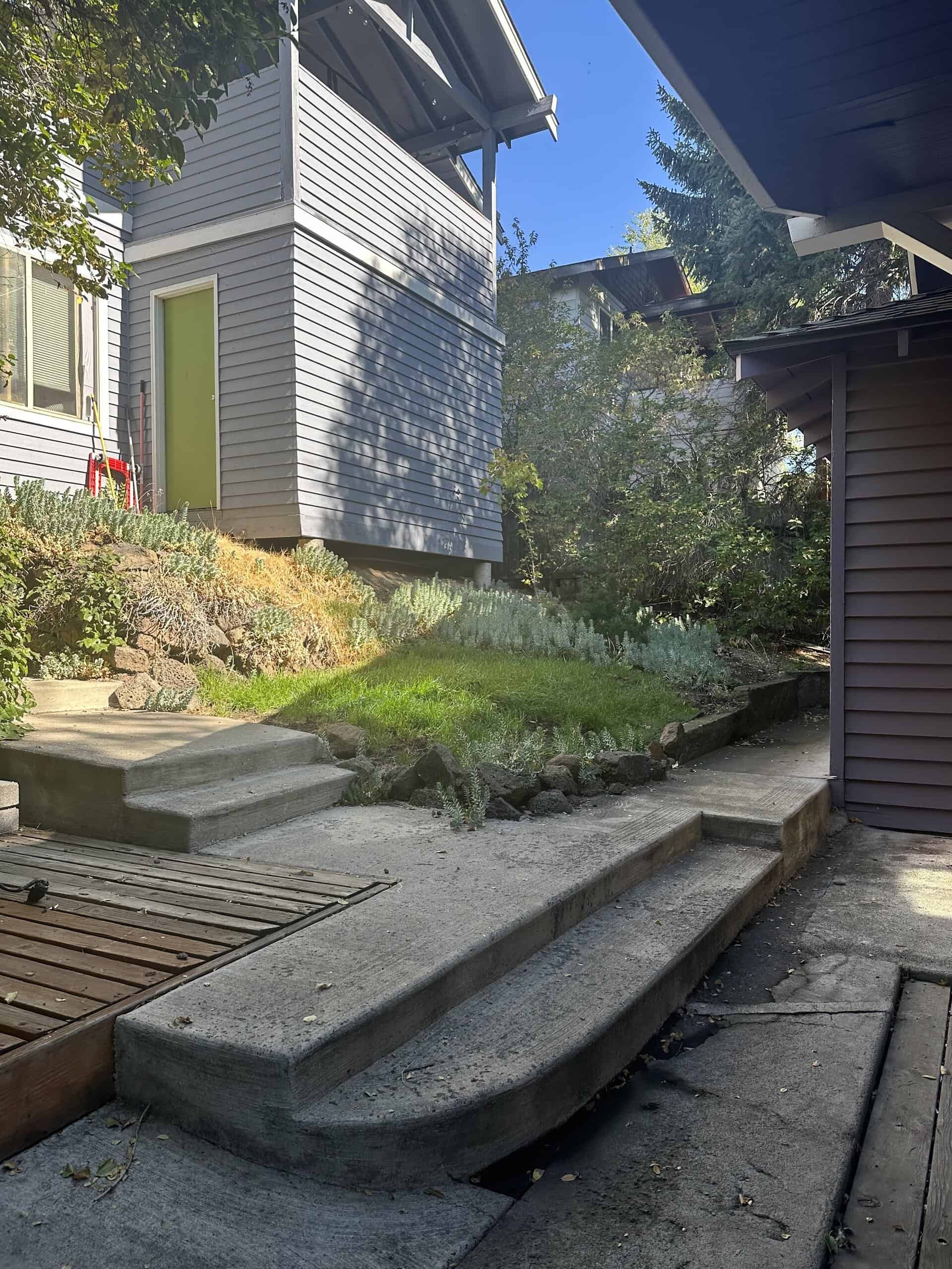 Concrete steps lead to a small grassy slope between gray-sided buildings, with shrubs and trees around it.