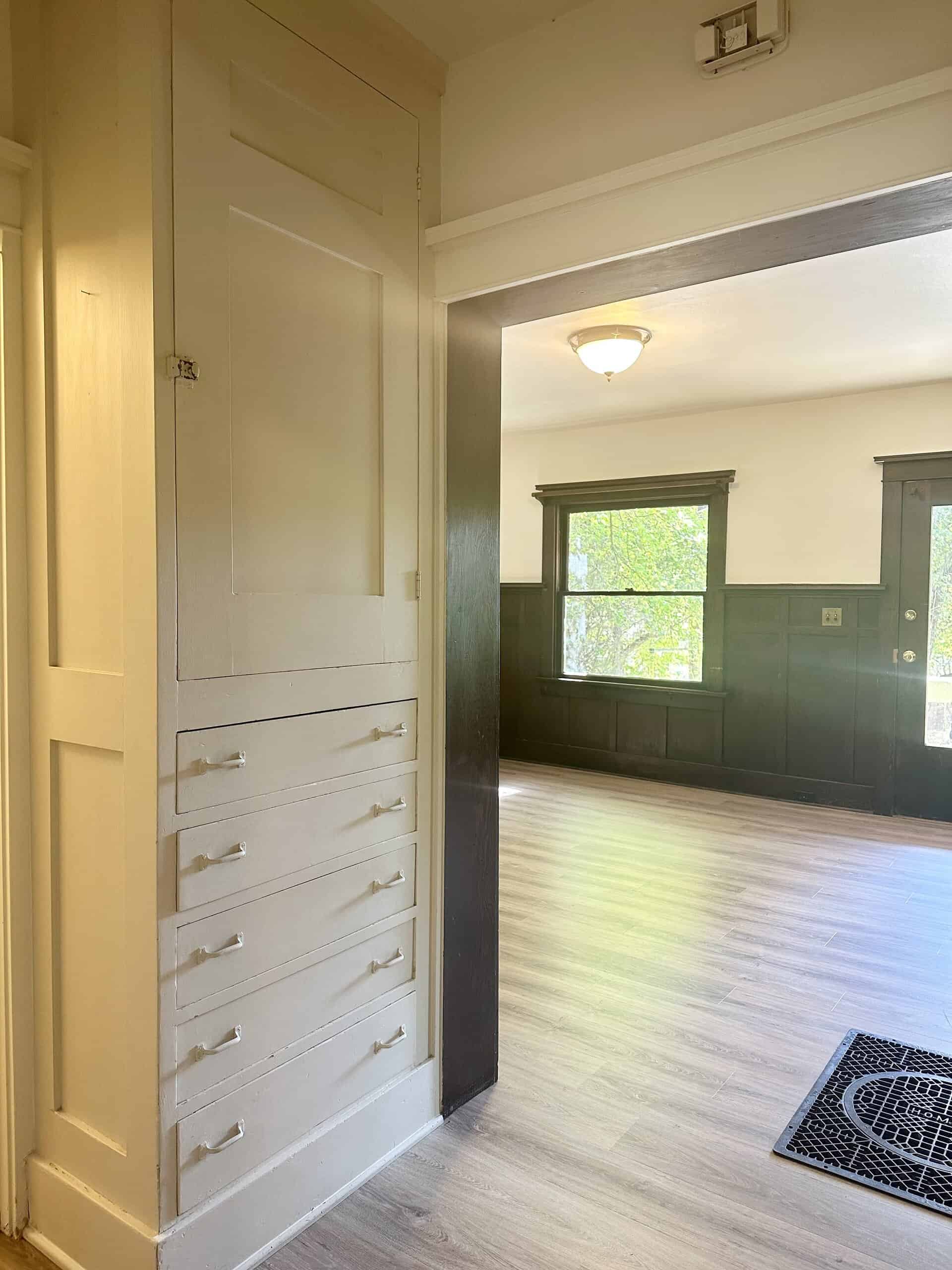 A doorway beside a built-in cabinet with several drawers opens onto a room with dark wall paneling, windows, and wood-pattern flooring.