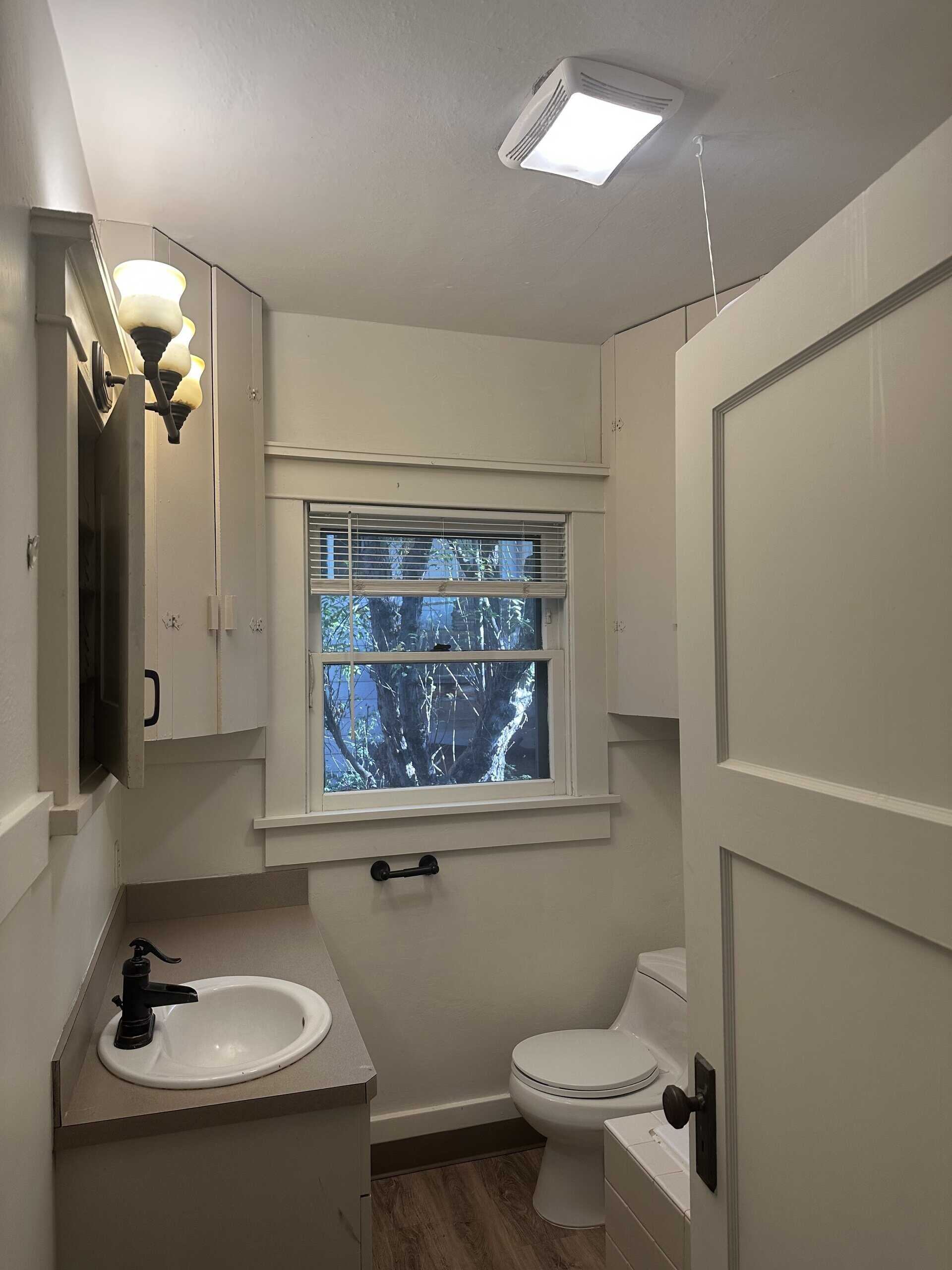 A bathroom with a sink, toilet, window with blinds, wall cabinets, and a lit ceiling fixture.